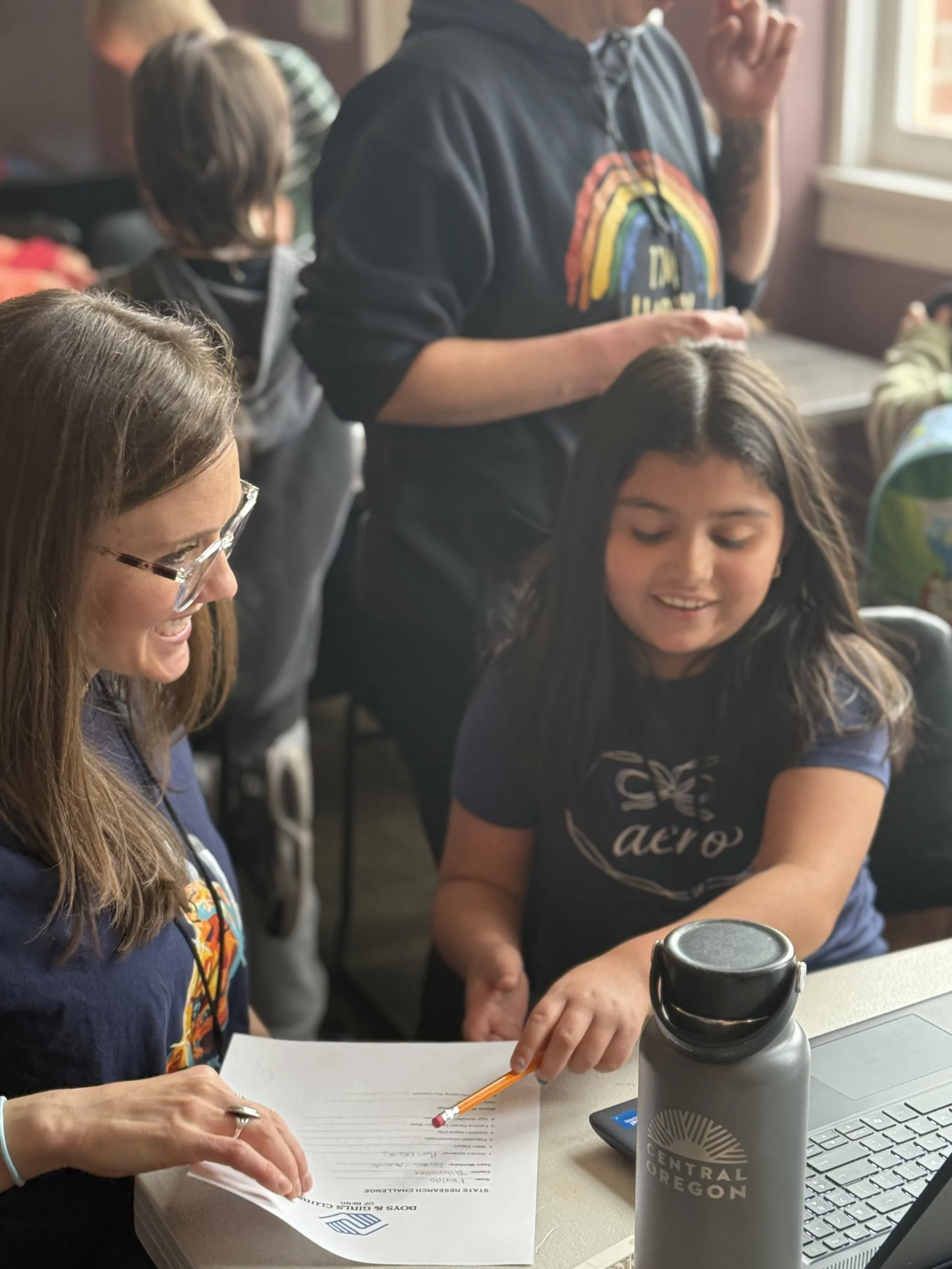 A group of people working together at a desk, with one woman smiling, taking notes, and a water bottle with 'Central Oregon' on it, a laptop, and papers.