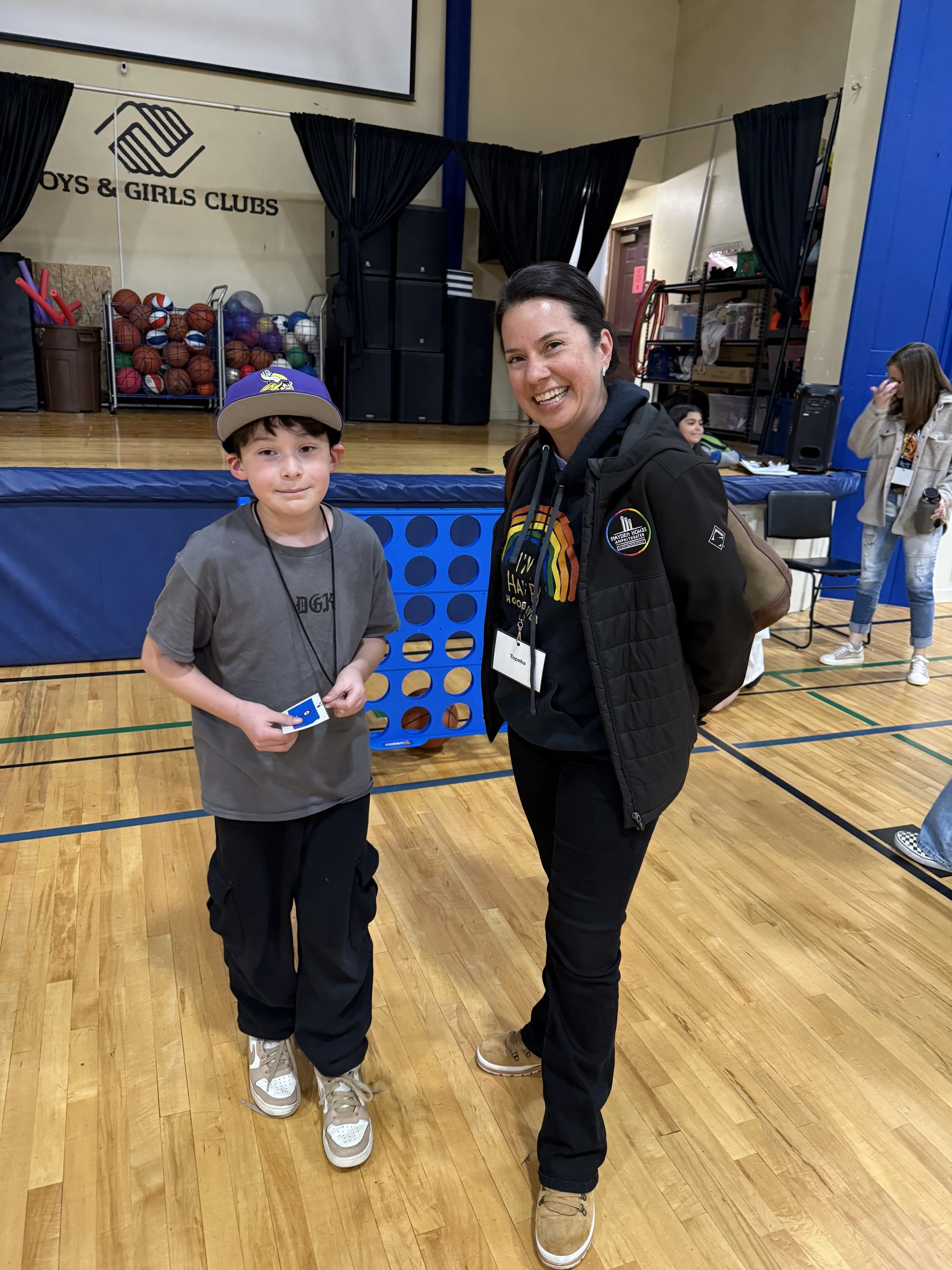 A woman and a young boy smiling and posing for a photo indoors at a Boys & Girls Clubs event. The background shows basketballs, sports equipment, and a stage with black curtains.