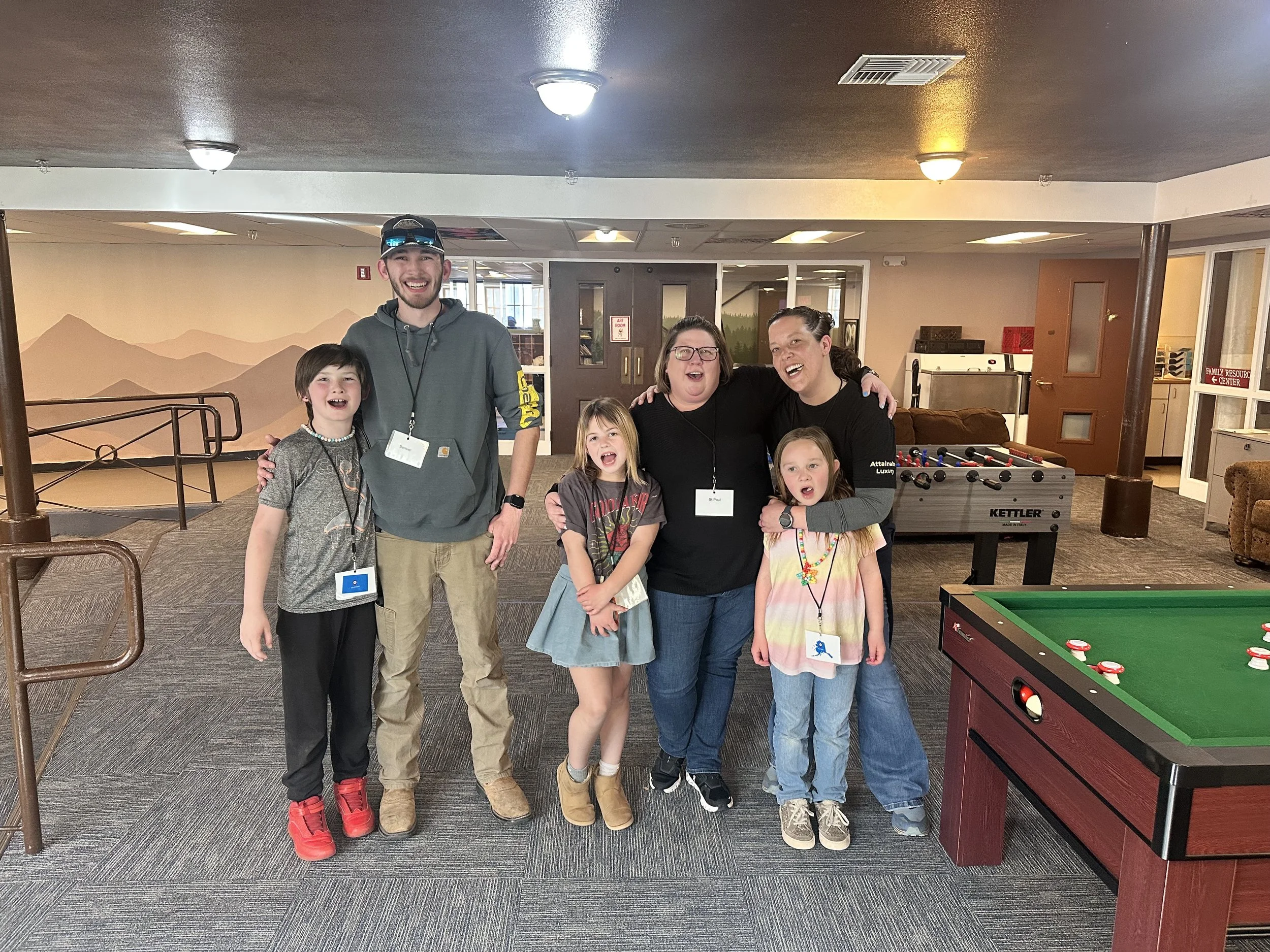 A group of six people, three adults and three children, standing together indoors, smiling and posing for a photo. There is a foosball table and a recreation area in the background.