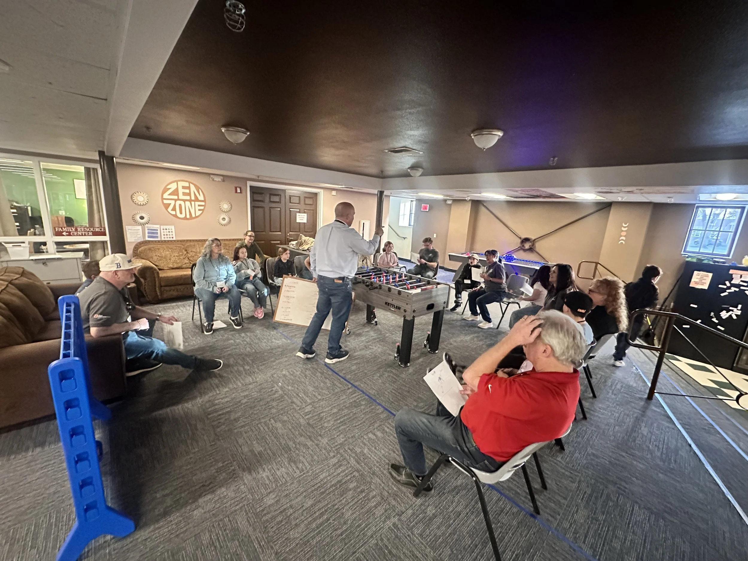 People gathered in a community room playing foosball and sitting on chairs, with a staff member instructing the group. The room has a sign that says 'ZEN ZONE' and a new game setup in the background.