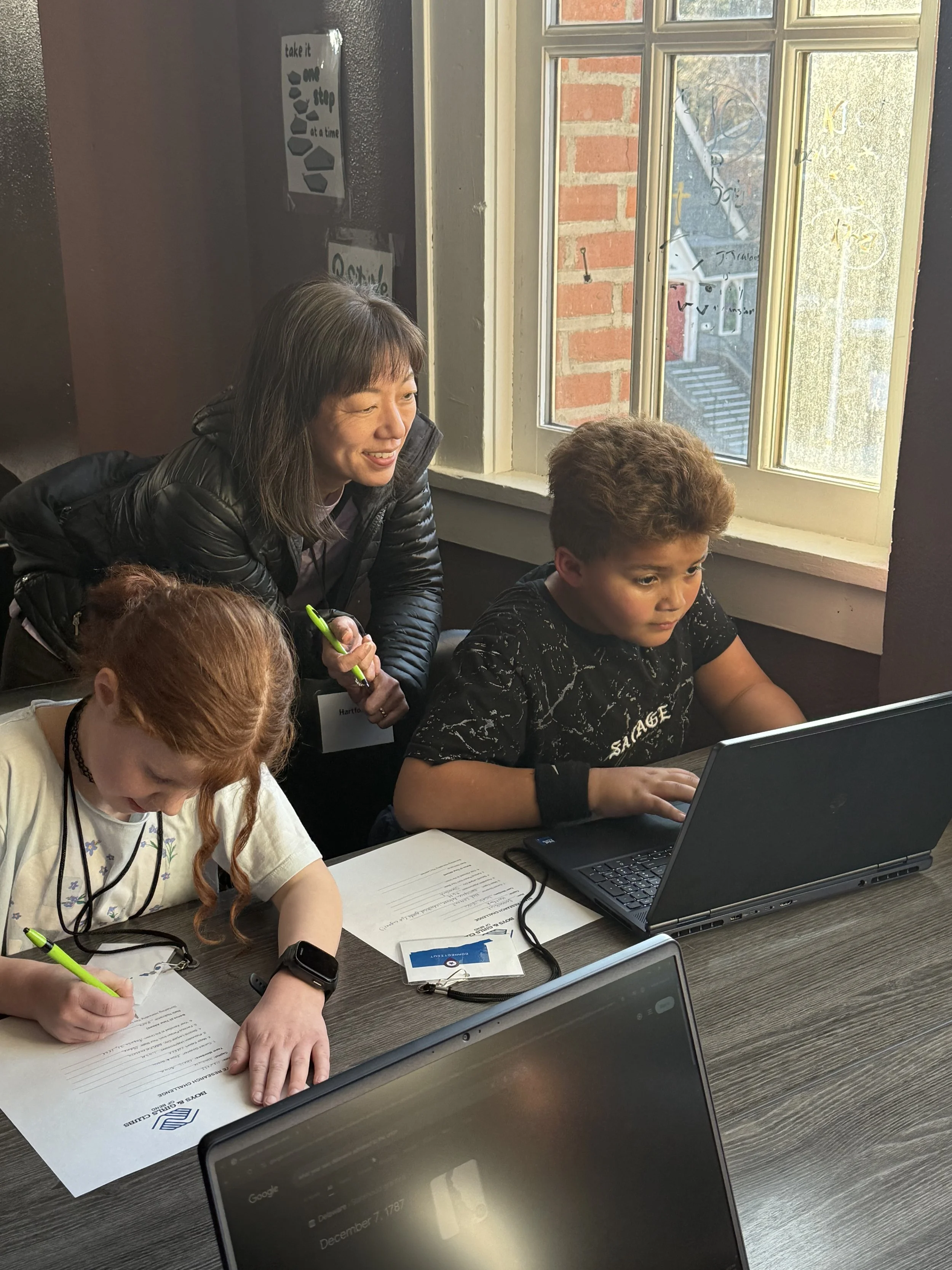 A woman with dark hair and a black jacket smiling while leaning over to look at a laptop screen, with a young boy with curly hair sitting and typing on a laptop. There are papers, a pen, and electronic devices on the table near them, with a window sh