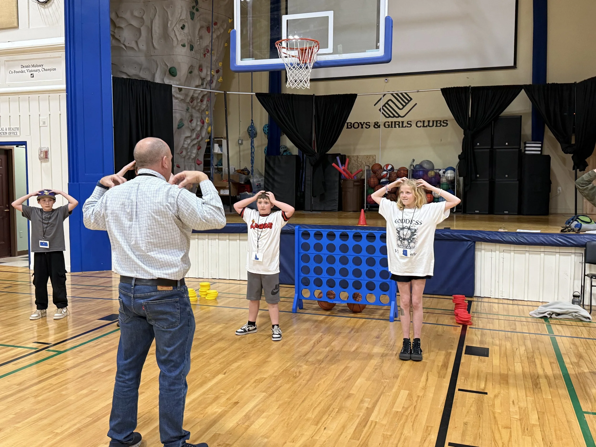 A group of children participating in a game of Connect Four, with a man supervising in a gymnasium decorated for a boys and girls club event. There is a basketball hoop overhead and a rock climbing wall in the background.