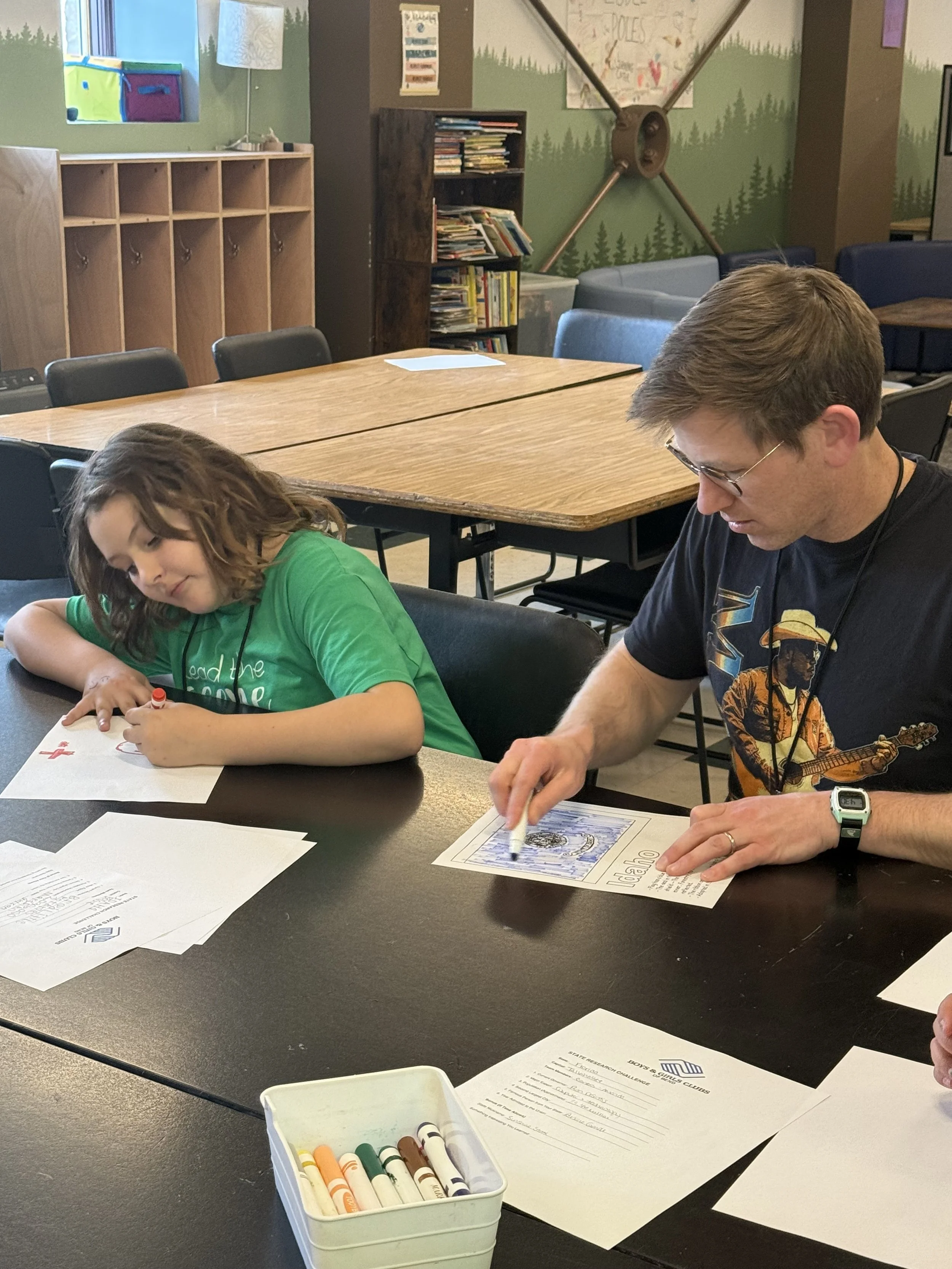 A young girl in a green shirt and an adult man with glasses and aT-shirt with a guitar design are sitting at a table, working on papers with printed text and coloring supplies, in a room with bookshelves, colorful storage bins, and a mural with trees