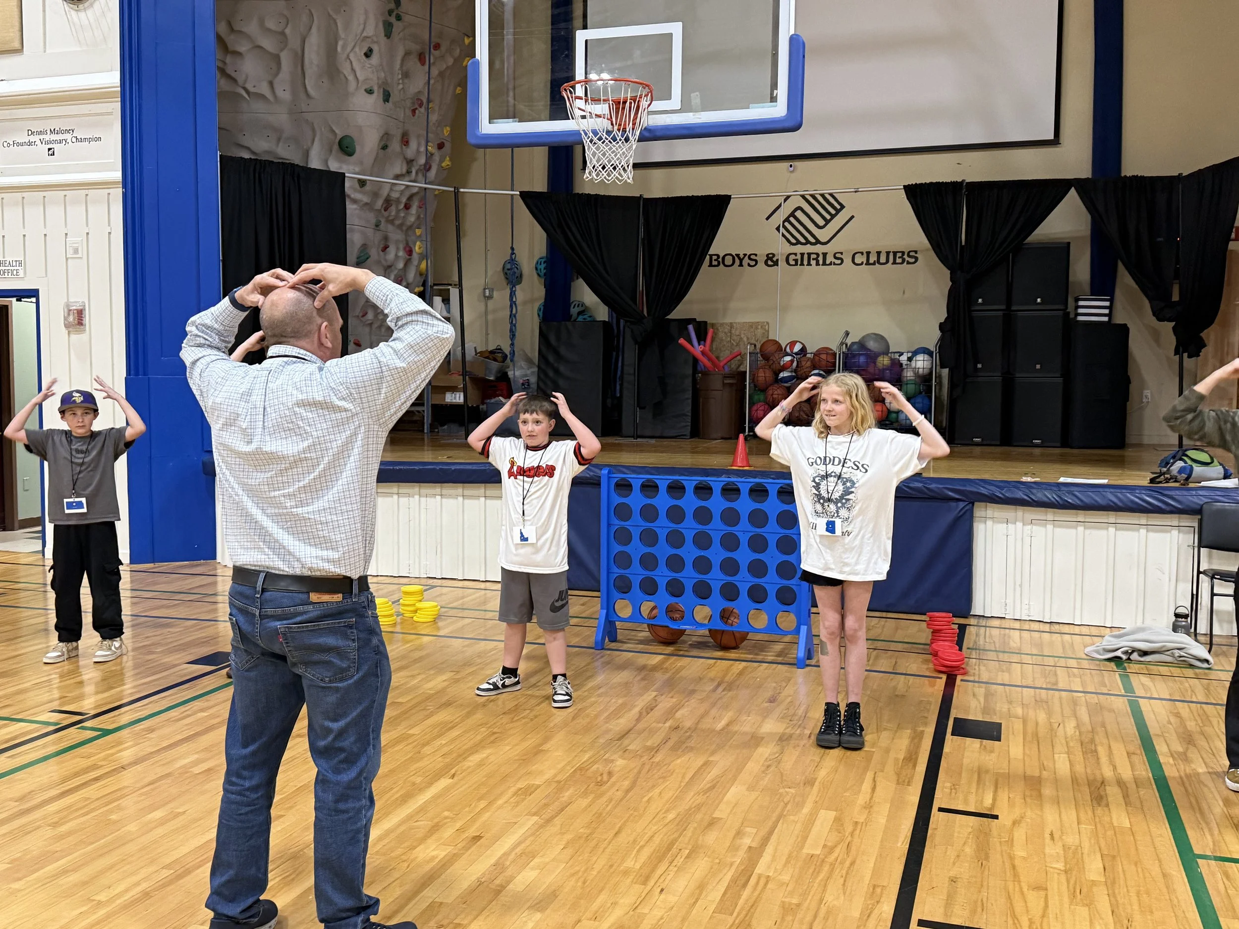 Children are participating in a game of Connect Four in a gymnasium, with a man supervising. Some children are standing with hands on their heads. The setting is inside a basketball gym with a basketball hoop and a rock climbing wall in the backgroun