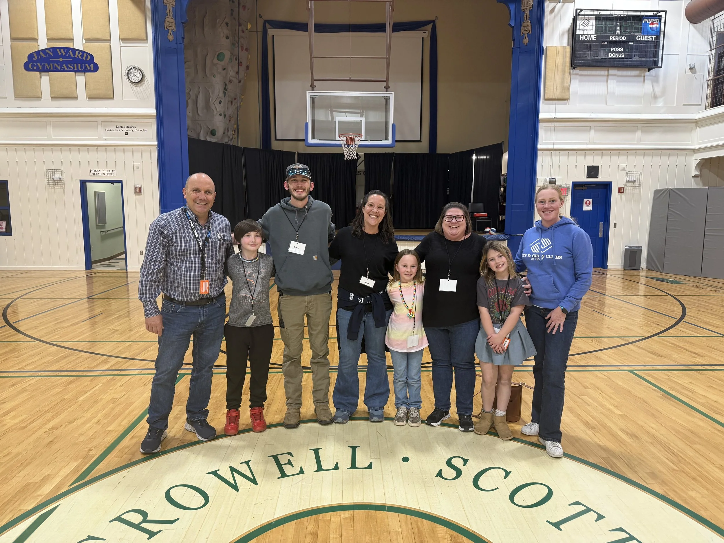A group of nine people, including children and adults, standing together inside a gymnasium, smiling for a photo. The gymnasium has wooden floors, a basketball hoop, black curtains, and a scoreboard. The group appears to be at a community or school e