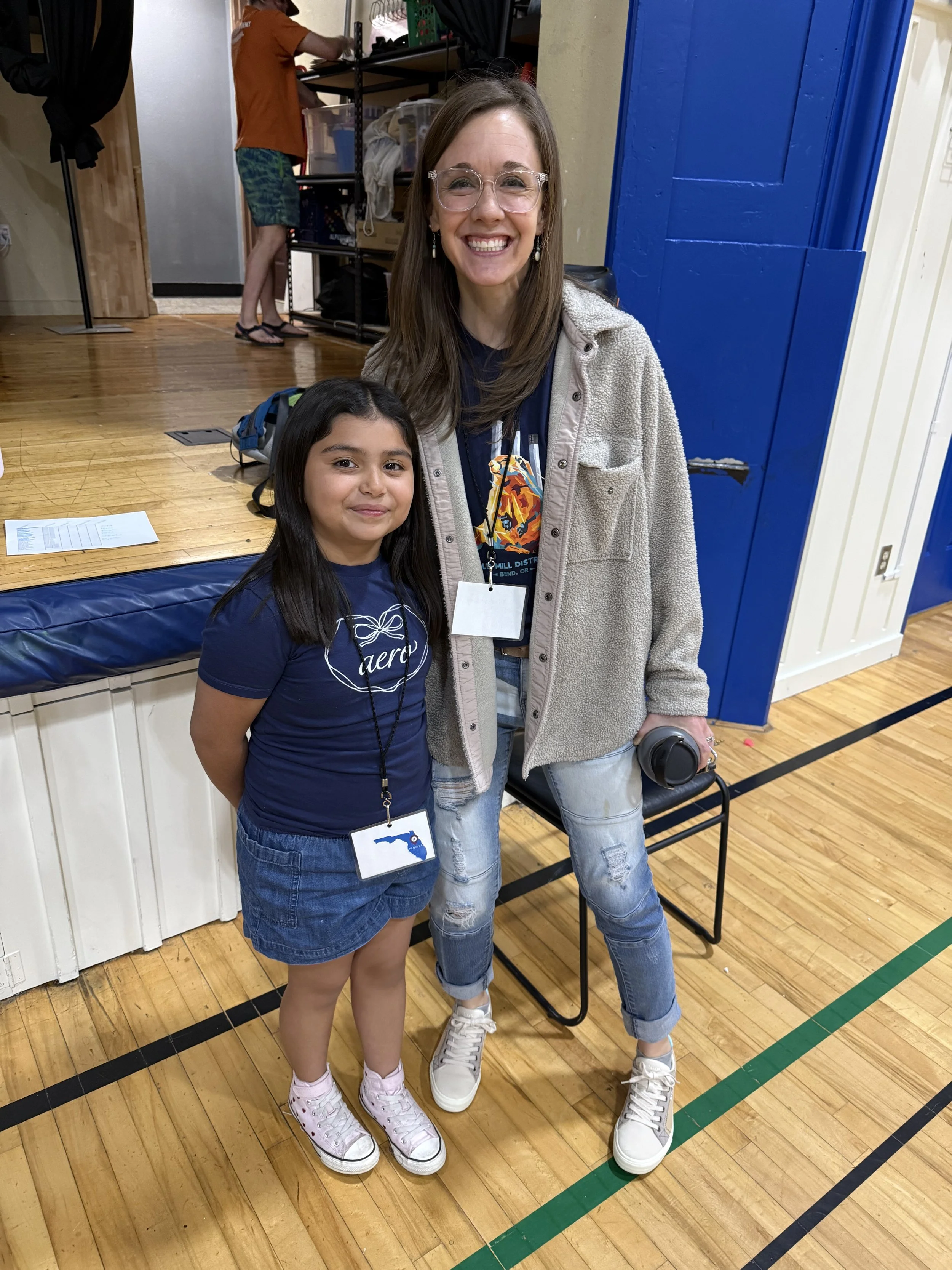 Two smiling girls standing on a wooden gymnasium floor. One girl has long dark hair, wearing a navy blue t-shirt with white sneakers, and a name tag with the shape of Florida. The other girl has long brown hair, wearing a beige jacket over a dark shi