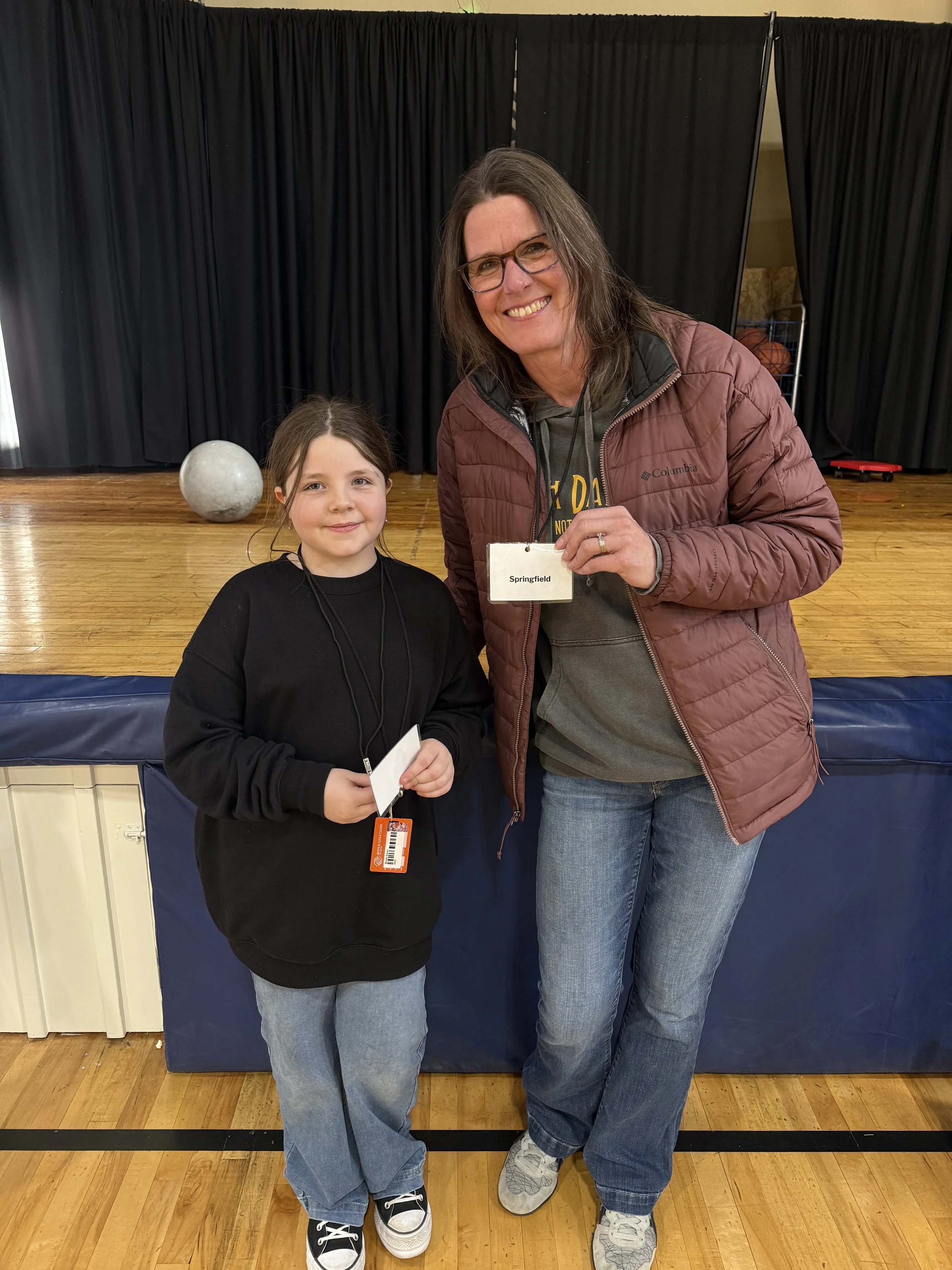 A woman with glasses and a brown jacket standing next to a young girl with long hair wearing a black sweatshirt inside a gymnasium or sports hall. The woman is holding a name tag that says "Springfield." They are both smiling, and there is a black cu