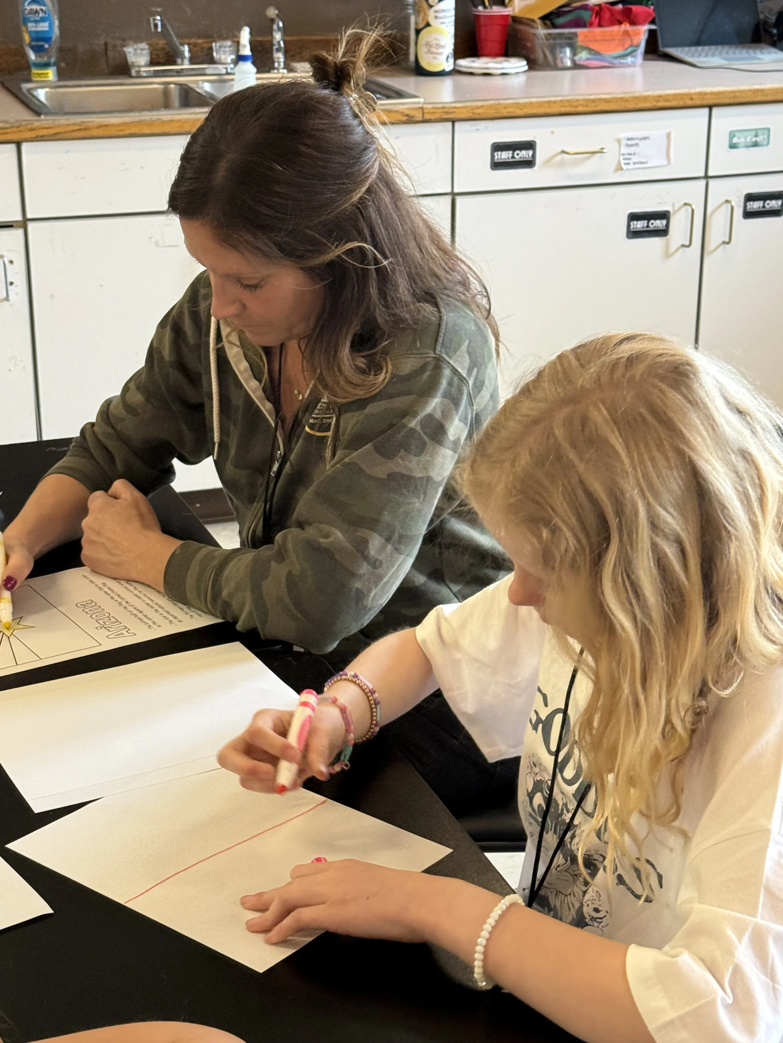 Two girls sitting at a table, coloring on sheets of paper with markers in a kitchen setting. The girl on the left has long, curly blonde hair, and the girl on the right has long, wavy brown hair. The kitchen has a sink, soap, and various items on the