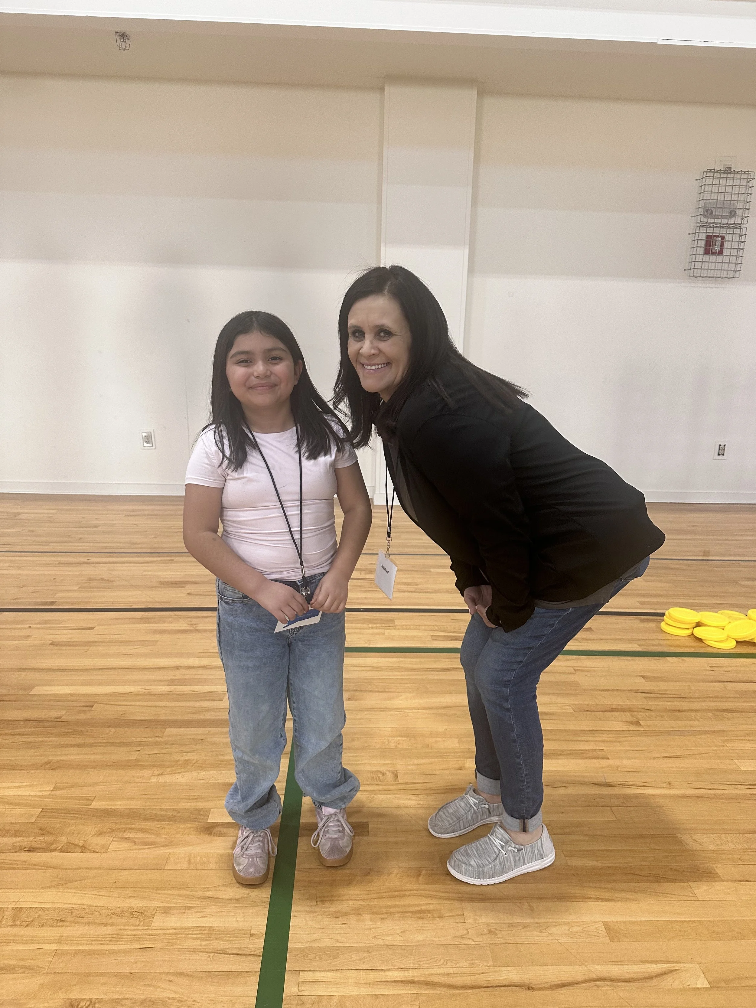 A woman and a young girl bend slightly forward, smiling, in a gymnasium with a wooden floor and a white wall.