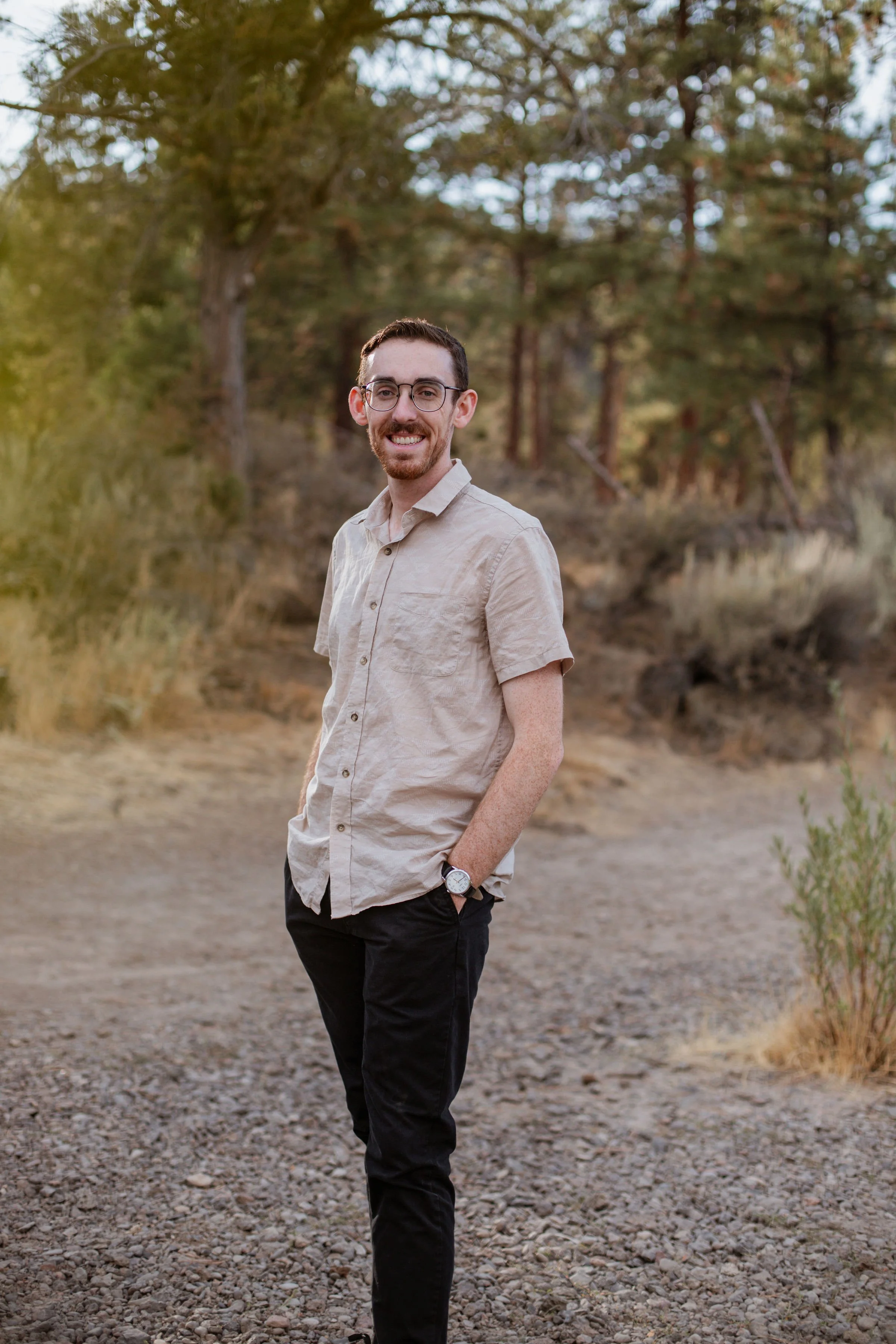 A man standing on a gravel path outdoors with trees in the background, smiling and wearing glasses, a beige short-sleeve shirt, black pants, and a watch.