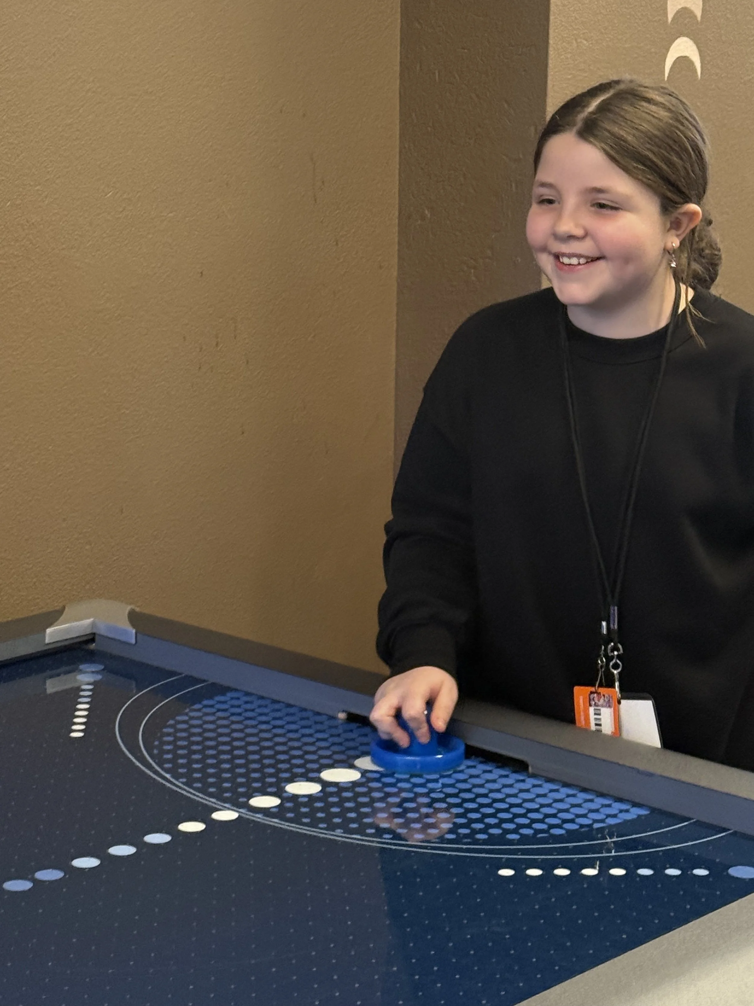 A young girl with brown hair smiling while playing a turntable game at an arcade or entertainment center.