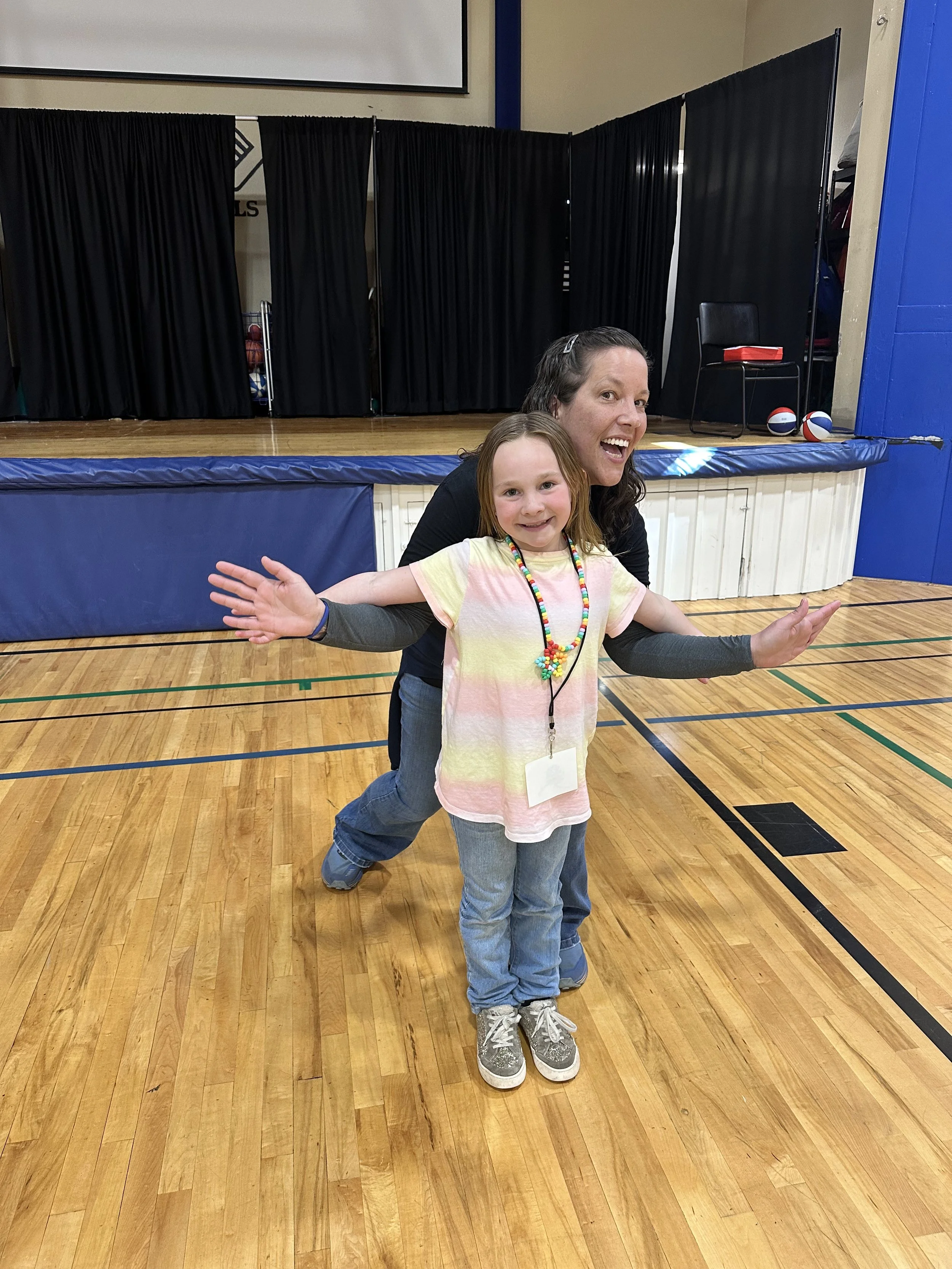 A woman and a young girl are posing playfully in a gymnasium, with the woman behind the girl, both wearing casual clothes and smiling. The girl has her arms outstretched and is wearing a rainbow-colored necklace. The background shows a small stage wi