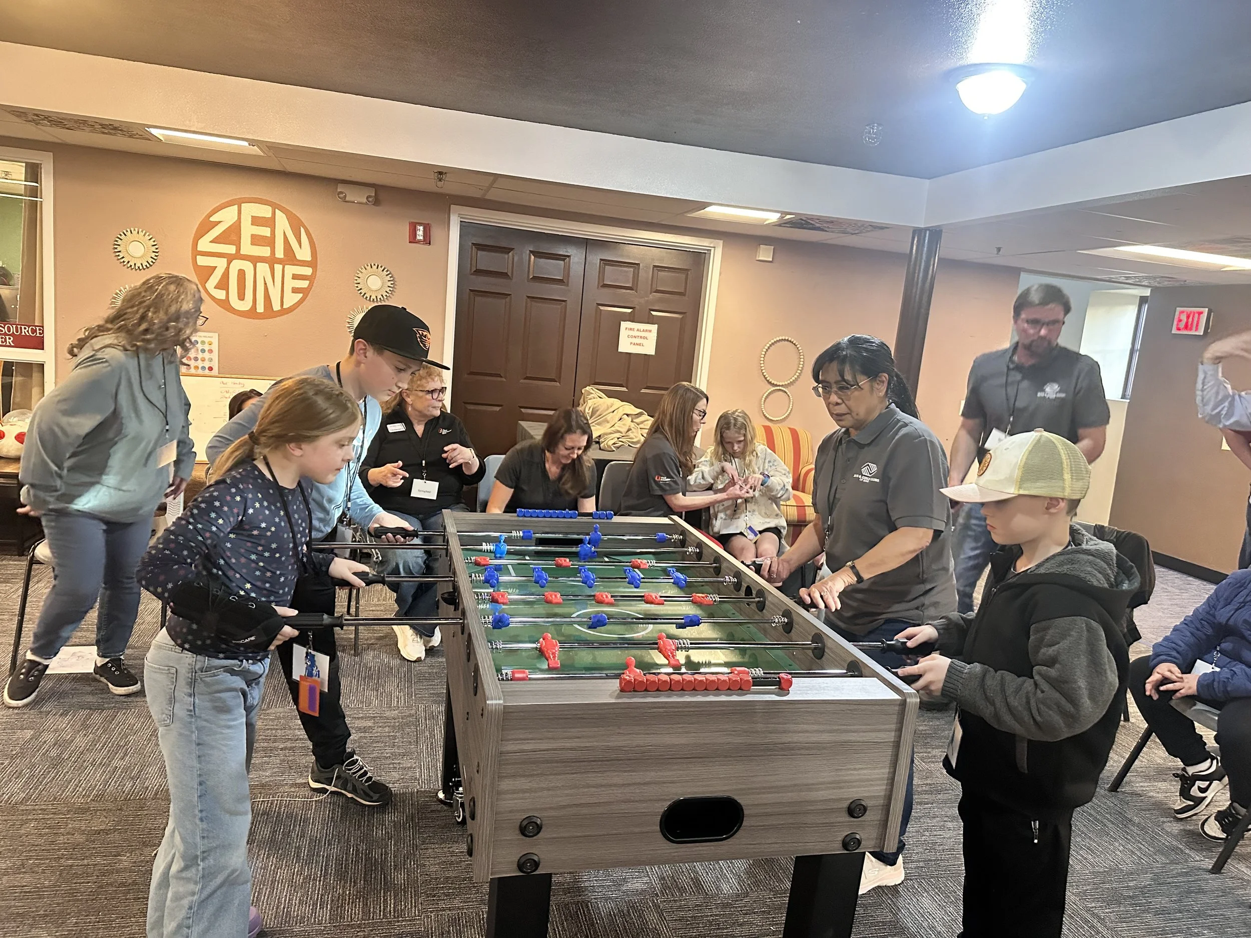 People playing a game of foosball in a public indoor space with a sign that says "ZEN ZONE" on the wall.