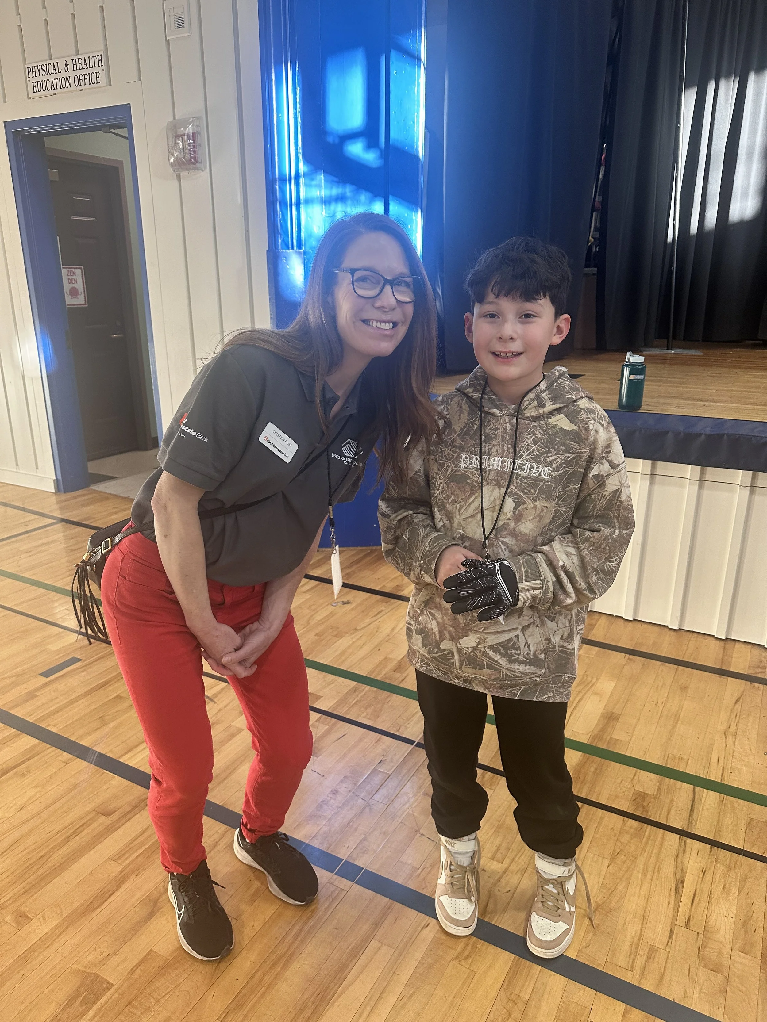 A woman with long hair, glasses, and a grey shirt is smiling and posing with a young boy who is wearing a camouflage hoodie, gloves, and rollerblades in a gymnasium. They are both smiling at the camera.