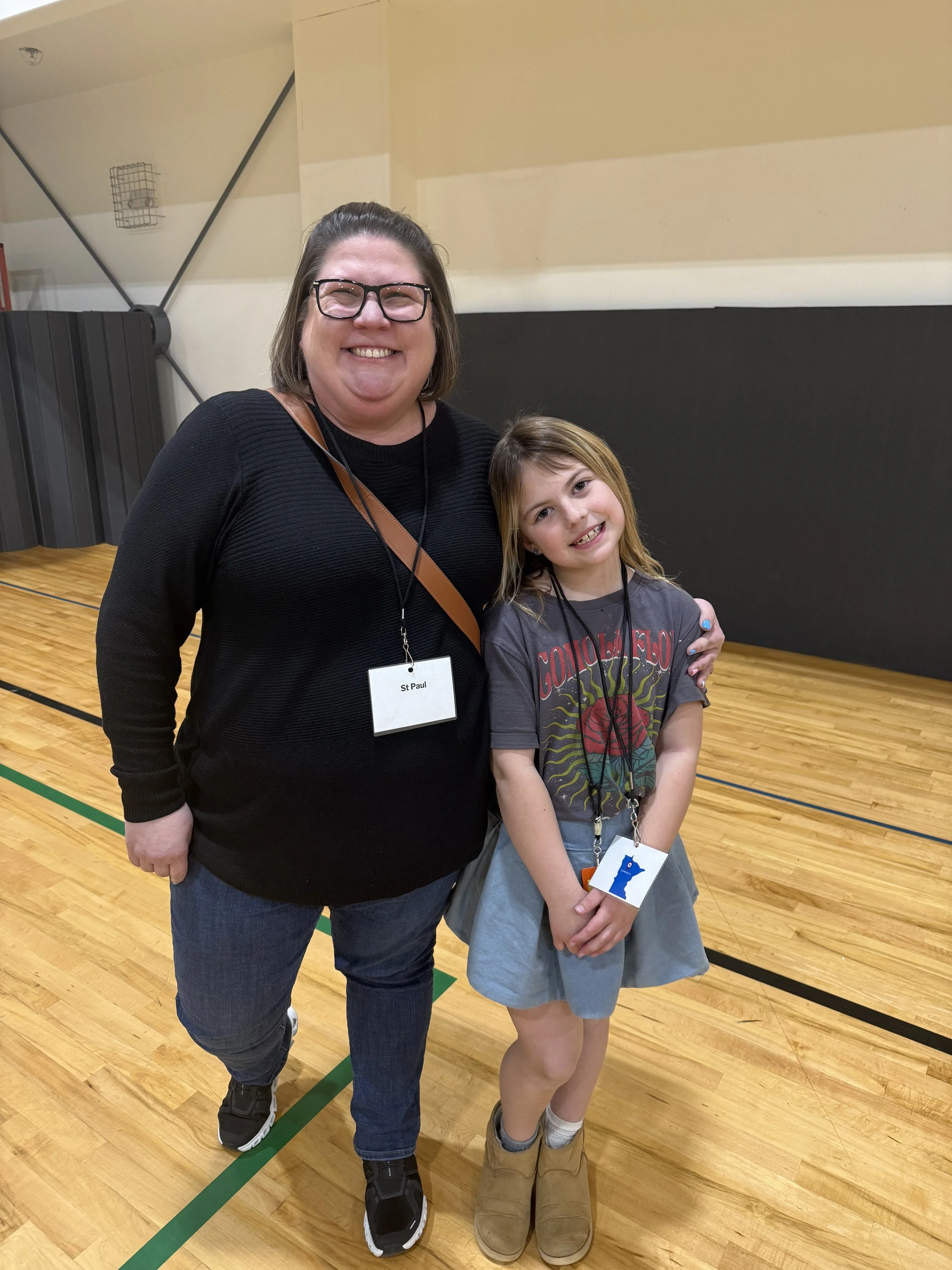 A woman and a young girl are smiling and posing together in a gymnasium with a wooden floor. The woman has glasses, shoulder-length brown hair, wearing a black top, blue jeans, and a name tag. The girl has long hair, wearing a graphic t-shirt, a gray