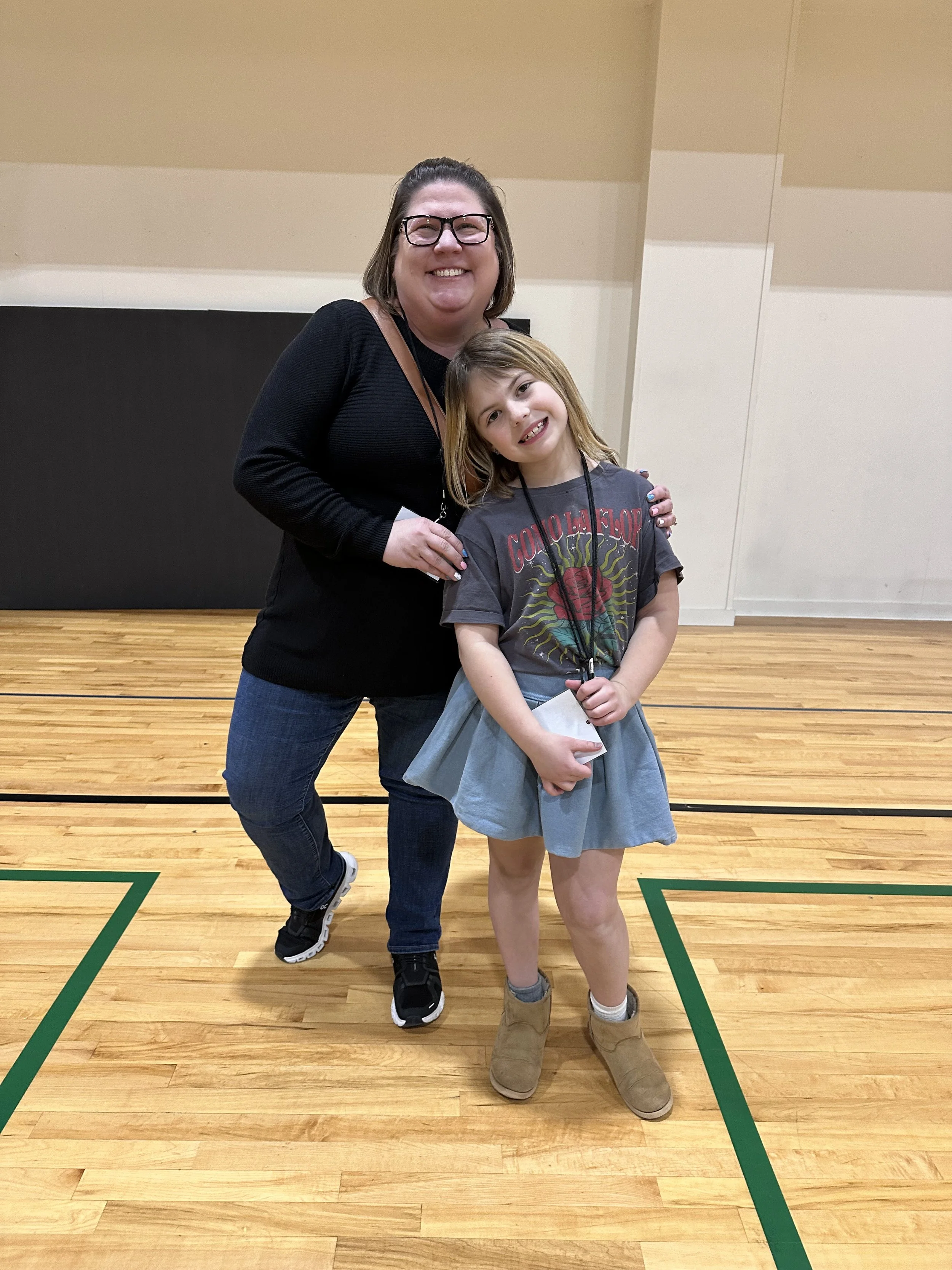 A woman with glasses and a black sweater smiling and hugging a young girl with long hair, a graphic t-shirt, a gray skirt, and tan boots, posing in a gymnasium with wood flooring and green lines.
