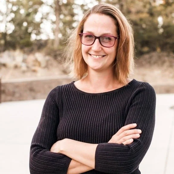 A smiling woman with shoulder-length light brown hair and red glasses, wearing a black long-sleeve ribbed sweater, standing outdoors with trees and a rocky landscape in the background.