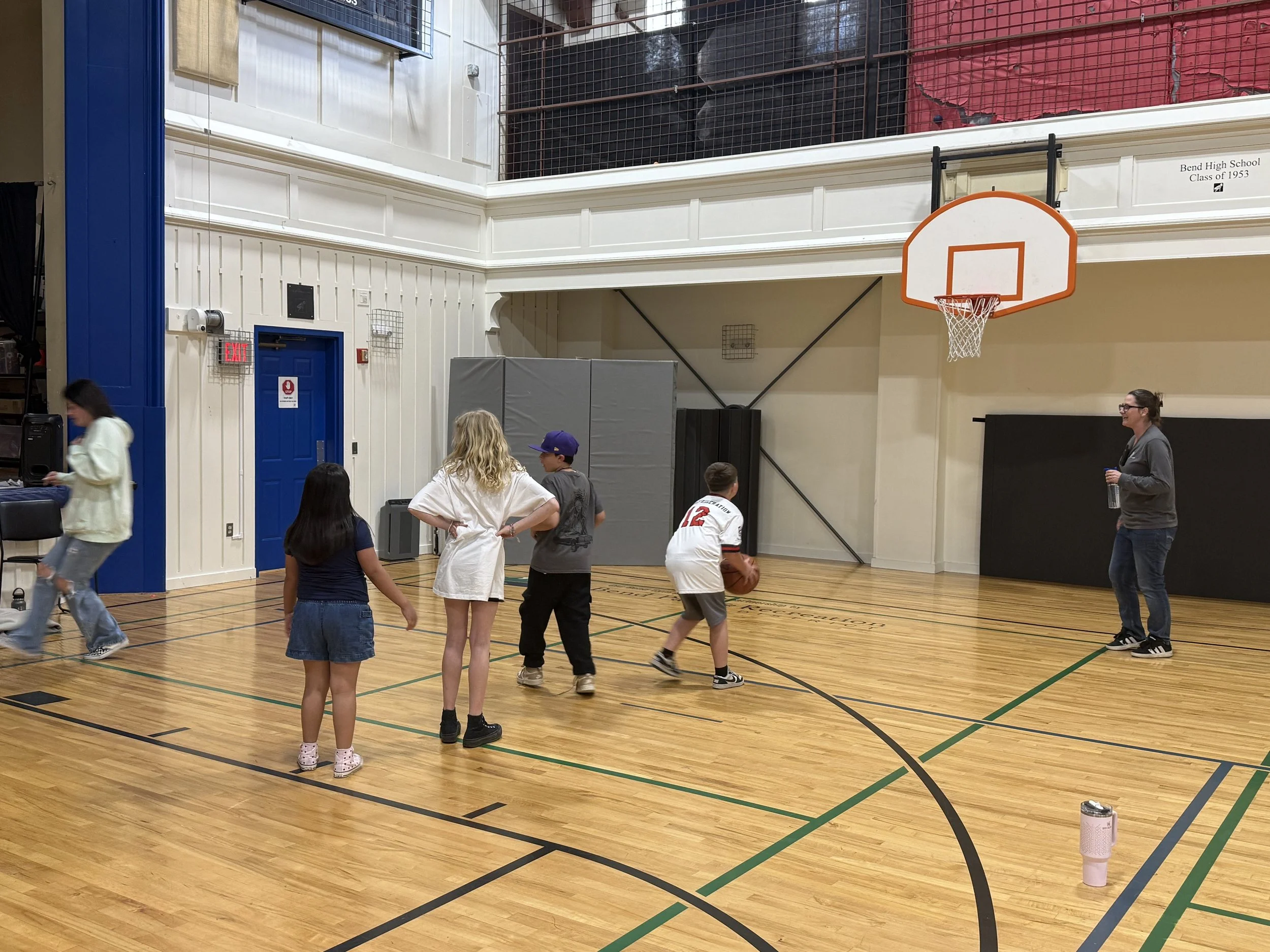 People playing basketball in an indoor gymnasium with wooden floors and a basketball hoop.