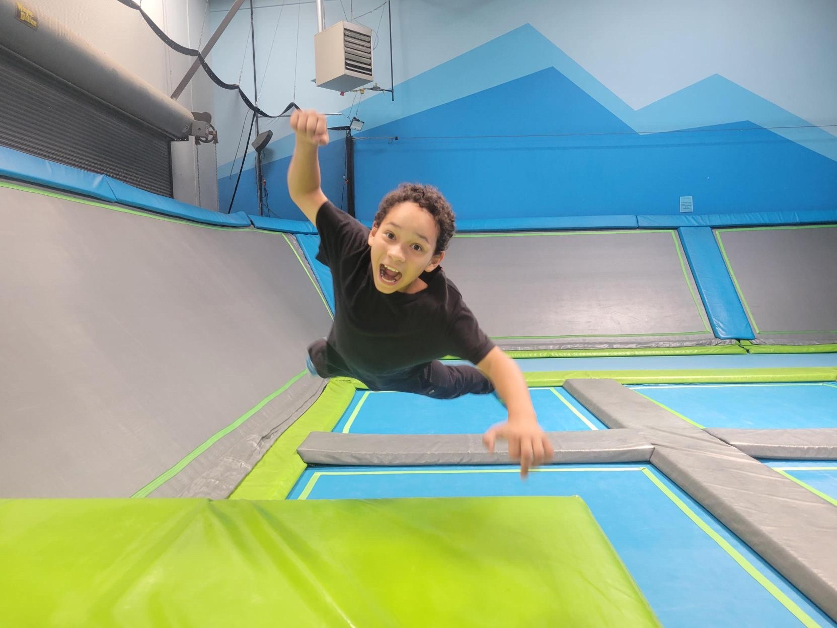 A young boy in a black shirt jumping on a trampoline in an indoor trampoline park, with a big smile and his mouth open.