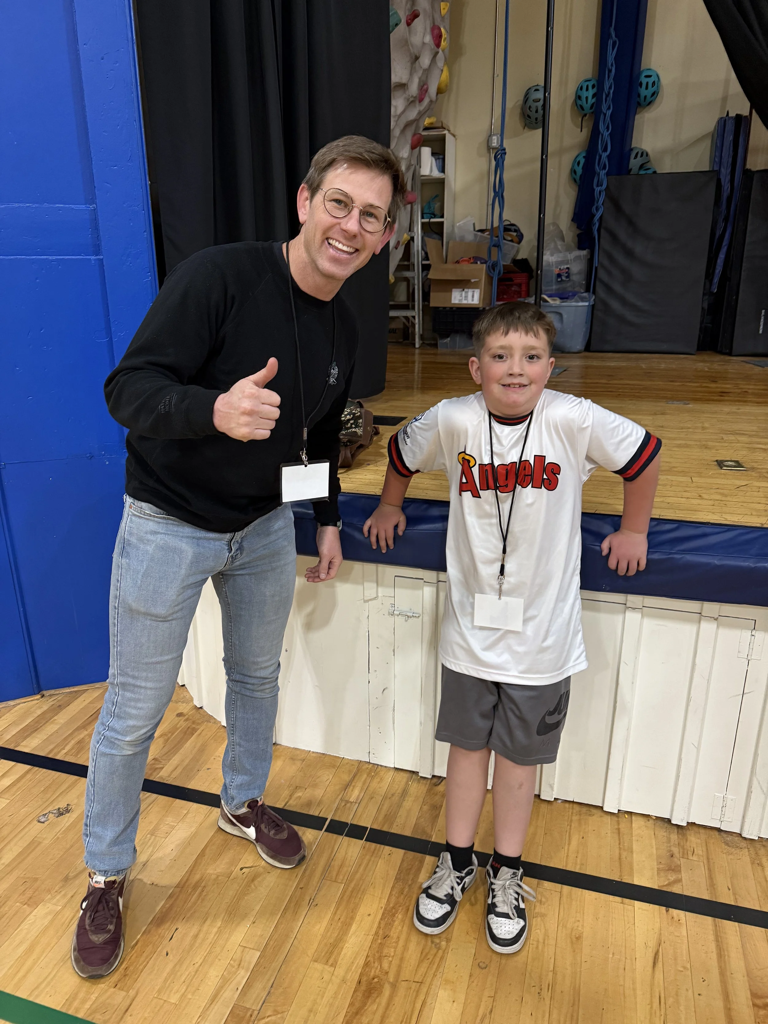 A man and a boy standing in a gymnasium, smiling for a photo. The man is giving a thumbs-up and the boy is wearing a sports jersey with 'Angels' written on it, both with medals around their necks.