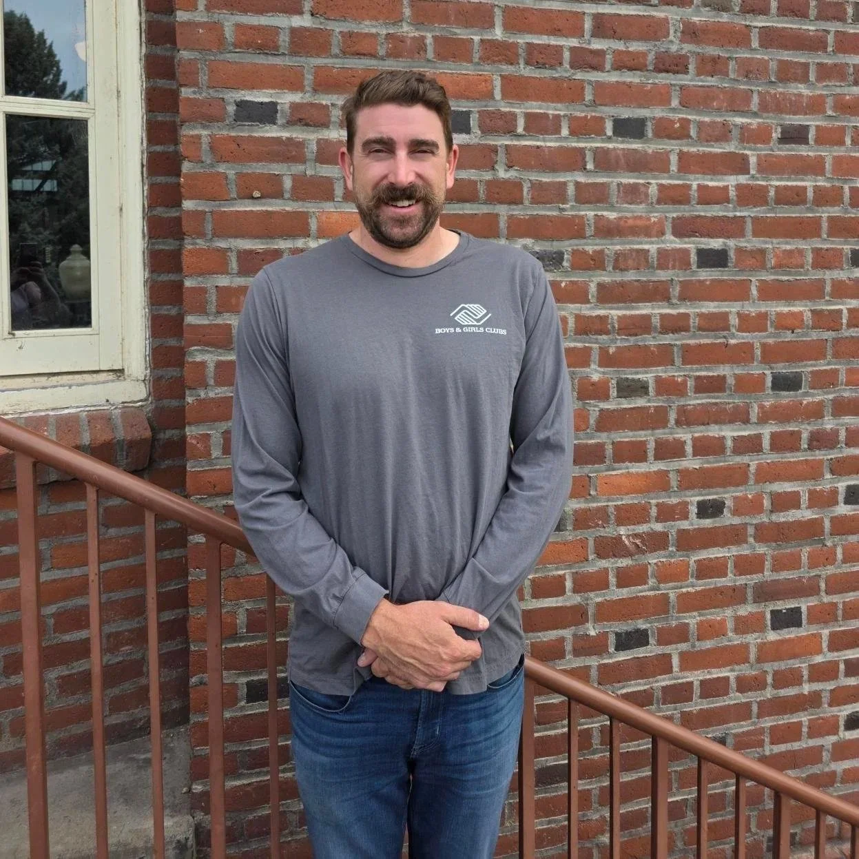 A man with a beard and short brown hair standing outdoors in front of a brick wall and window, wearing a gray long-sleeve shirt with a logo that says 'Boys & Girls Clubs' and blue jeans, smiling at the camera.