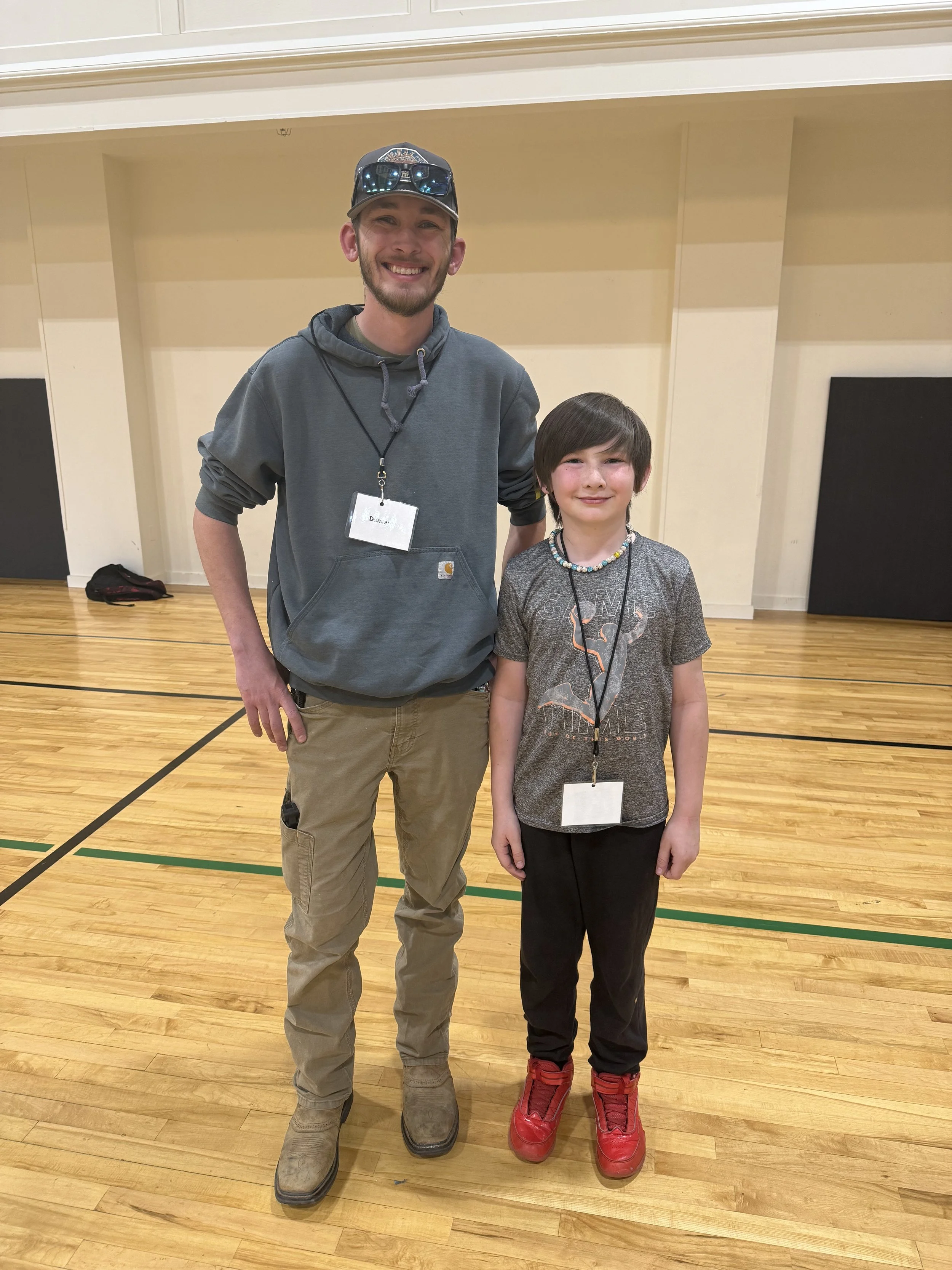 A young man and a young boy standing inside a gymnasium, smiling at the camera. The man is wearing a gray hoodie, khaki pants, and a cap with sunglasses on top, with a name tag around his neck. The boy is wearing a gray T-shirt with graphics, black p