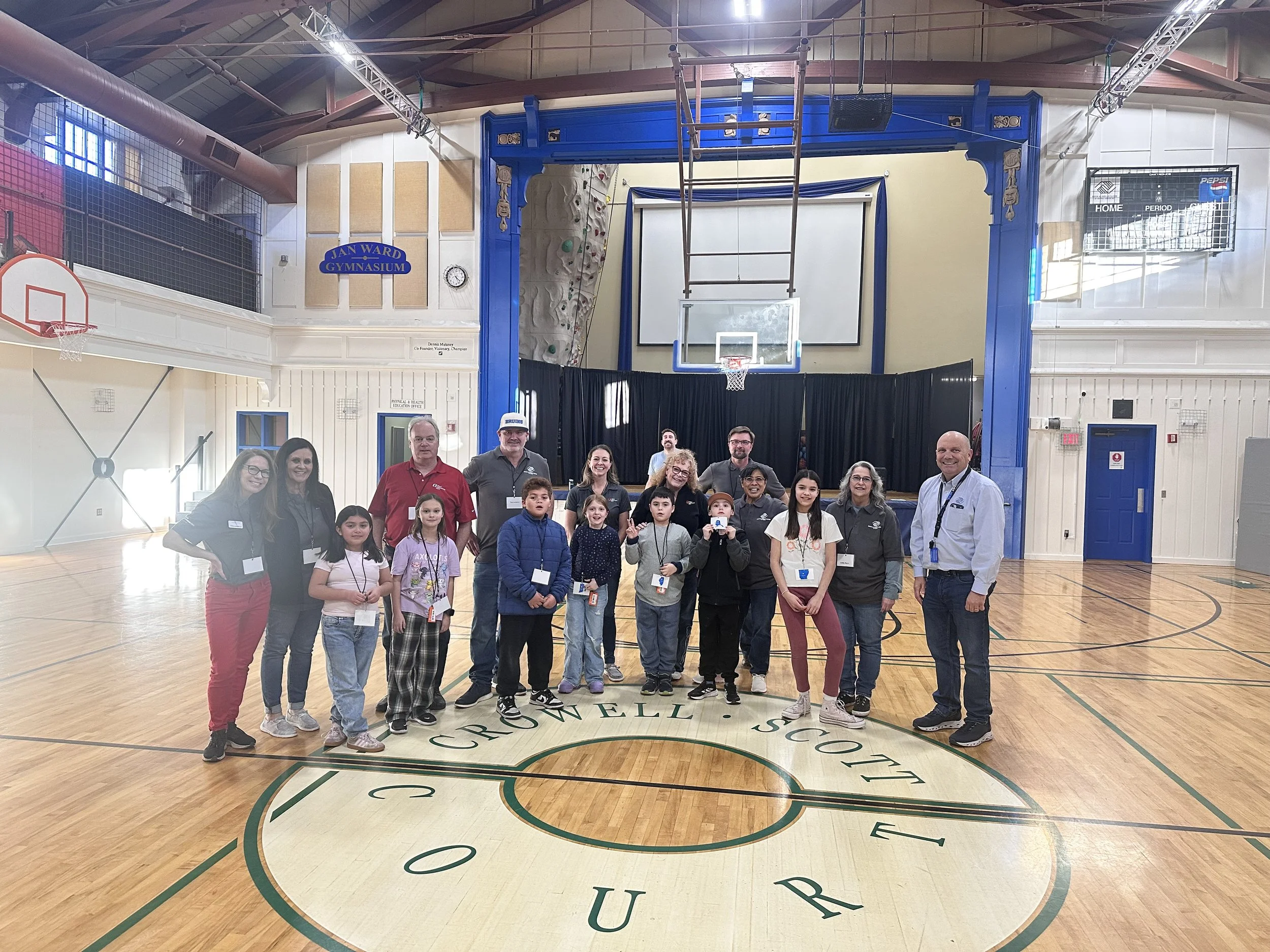 A group of children and adults posing for a photo inside a gymnasium, with a basketball hoop, climbing wall, and scoreboard visible in the background.