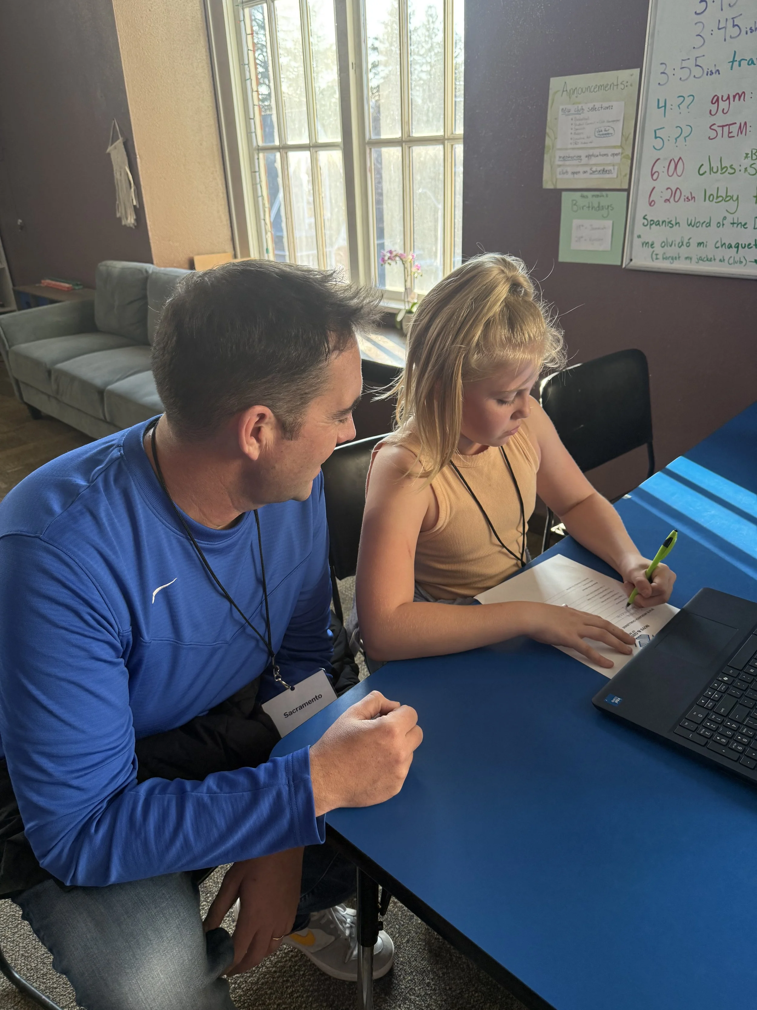 A man and a young girl are sitting at a blue table, looking at a book together. The man is wearing a blue shirt, and the girl is wearing a tan sleeveless top. They are indoors near a window with sunlight coming through, with a dark purple and beige w