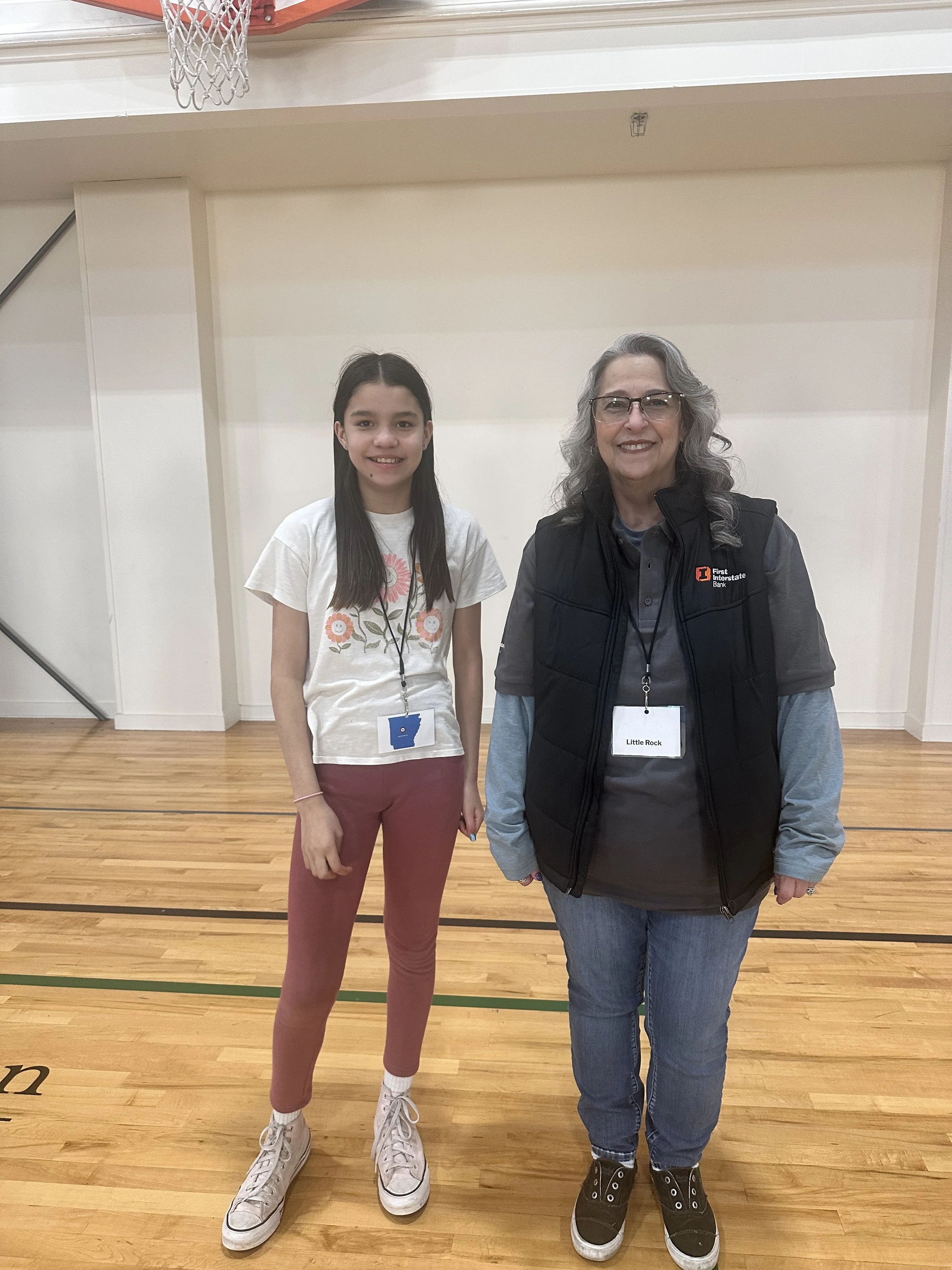 A young girl and an older woman standing side by side in a gymnasium, both smiling at the camera. The girl has long dark hair, is wearing a white T-shirt with a flower design, maroon pants, and white sneakers. The woman has curly gray hair, glasses, 