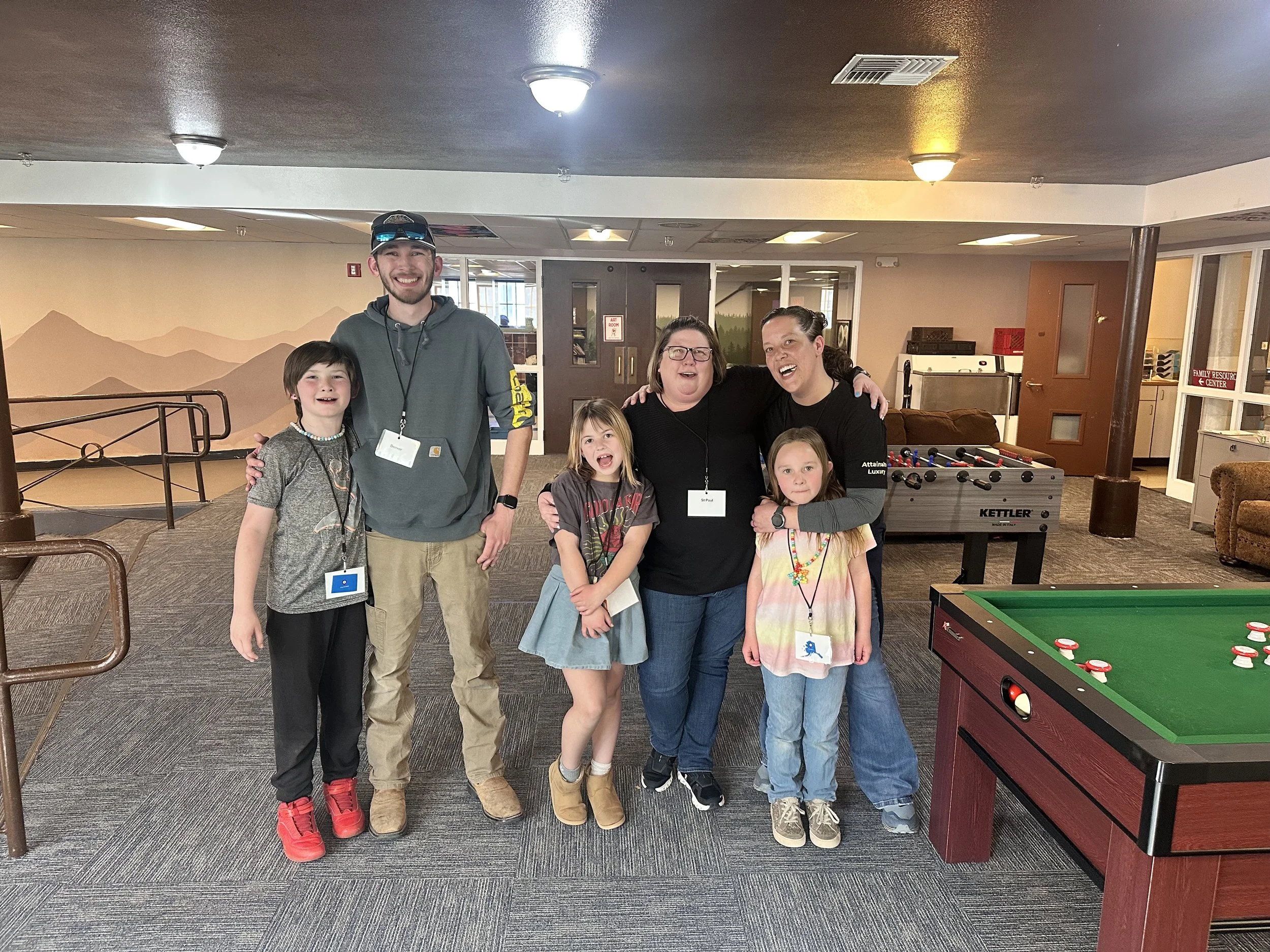 Group of six people, including children and adults, posing together indoors near a foosball table, with a background mural of mountains and a game room setting.