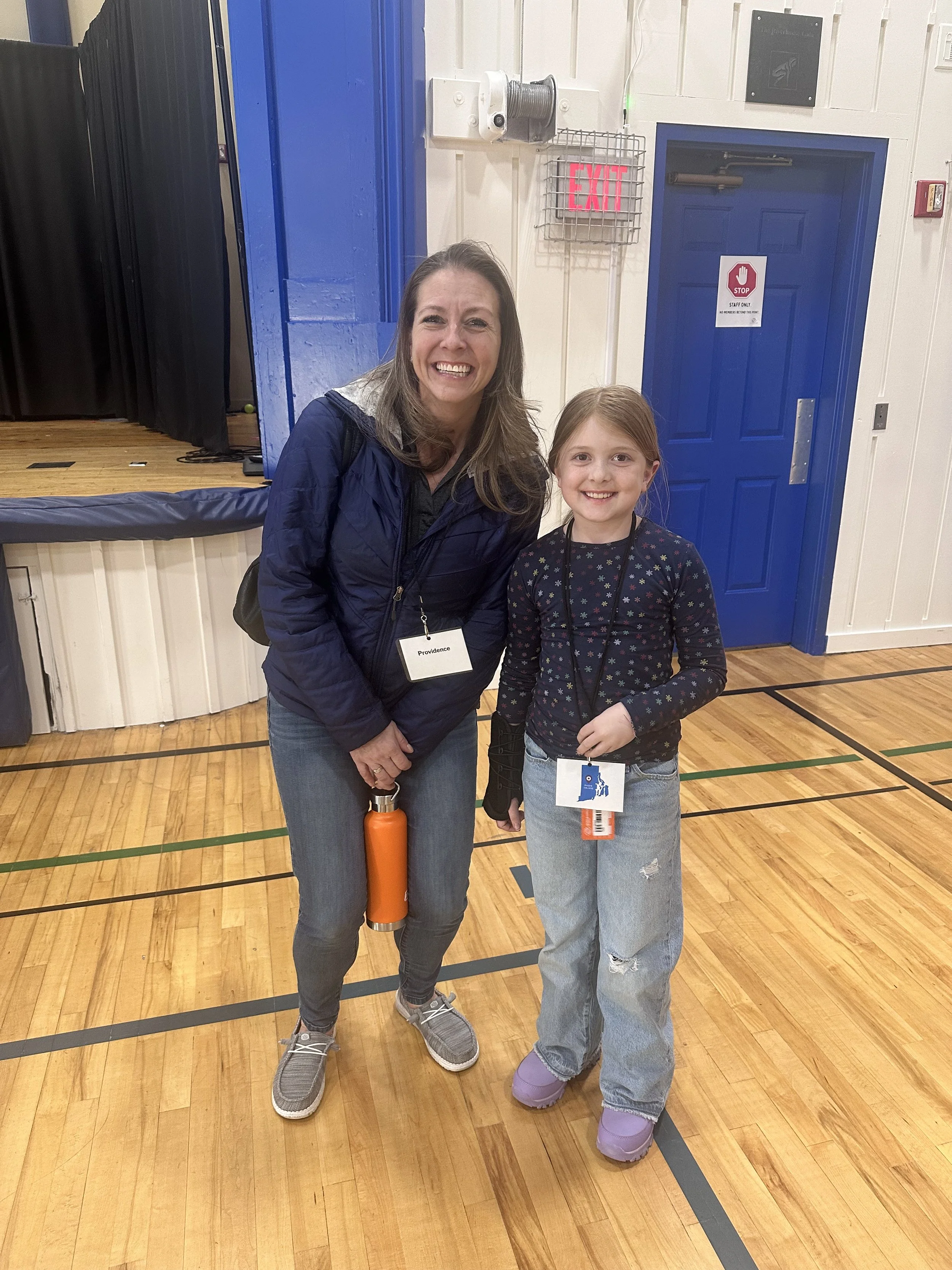 A woman and a young girl smiling and posing together in a gymnasium. The woman is holding a water bottle and wearing a dark jacket with a backpack. The girl is wearing oversized purple boots, distressed jeans, and a dark long-sleeve shirt with floral