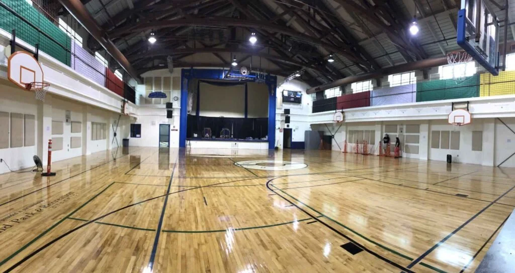 Empty indoor basketball court with a stage at the far end, basketball hoops on both sides, and hardwood flooring.