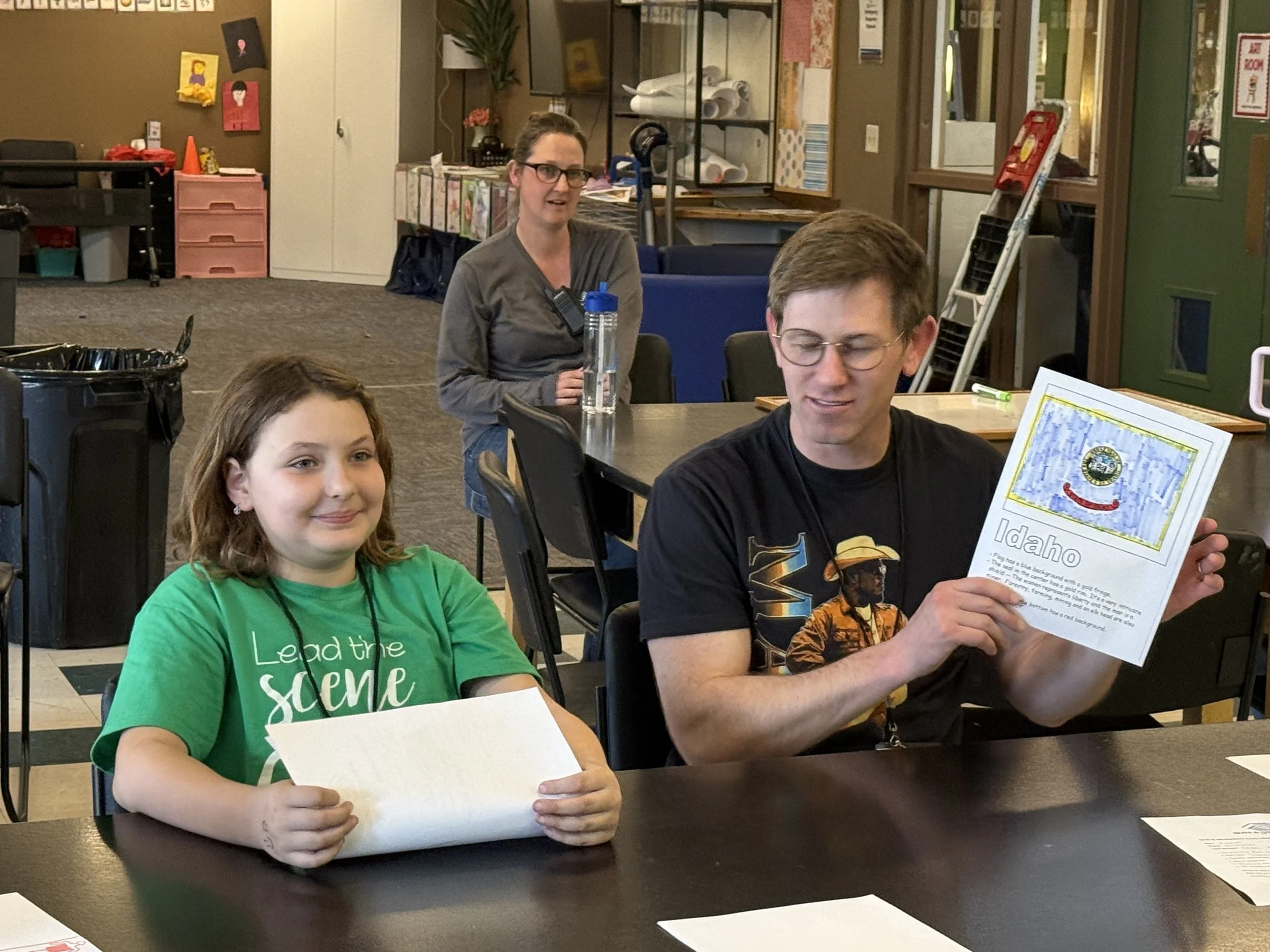 A young girl in a green t-shirt smiling at the camera, sitting at a table with a white paper in her hands. Behind her, a woman with glasses and a gray shirt stands, looking in the direction of the girl. The setting appears to be an indoor room with v