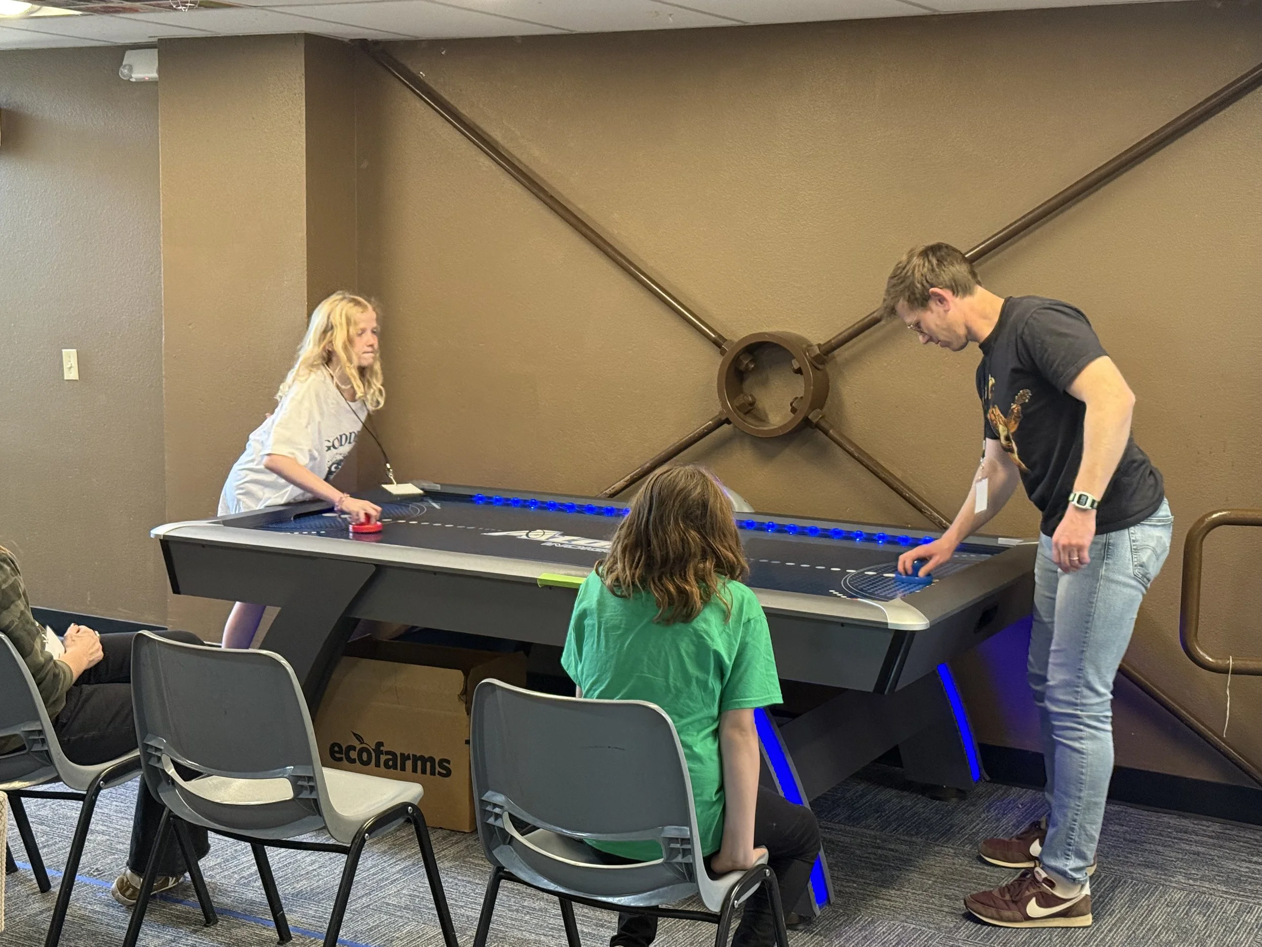 A group of kids and adults playing air hockey in a room with gray walls and carpet.