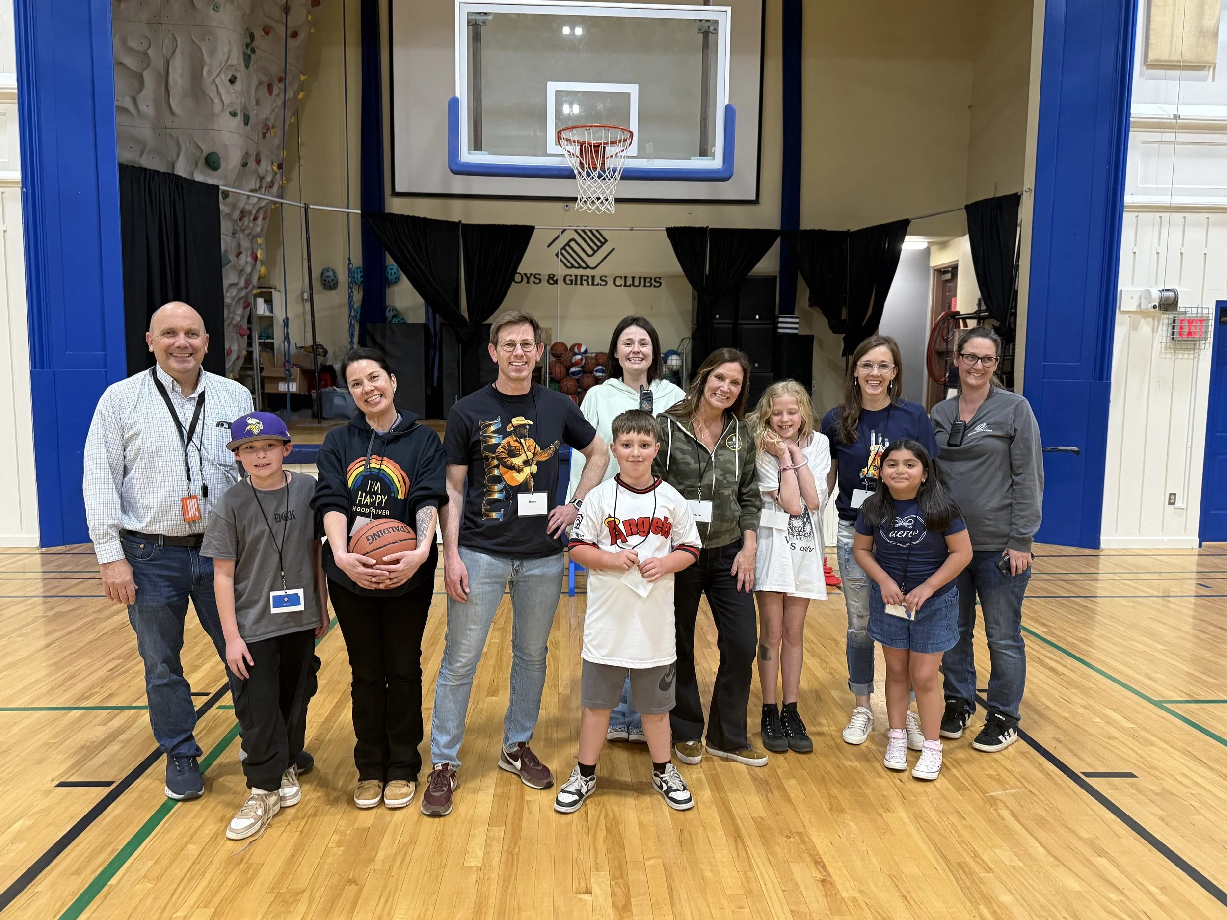 A group of adults and children smiling and posing for a photo in a gymnasium with a basketball hoop and climbing wall in the background. The group includes three men, four women, and four children, some holding basketballs. The gym has a wooden floor