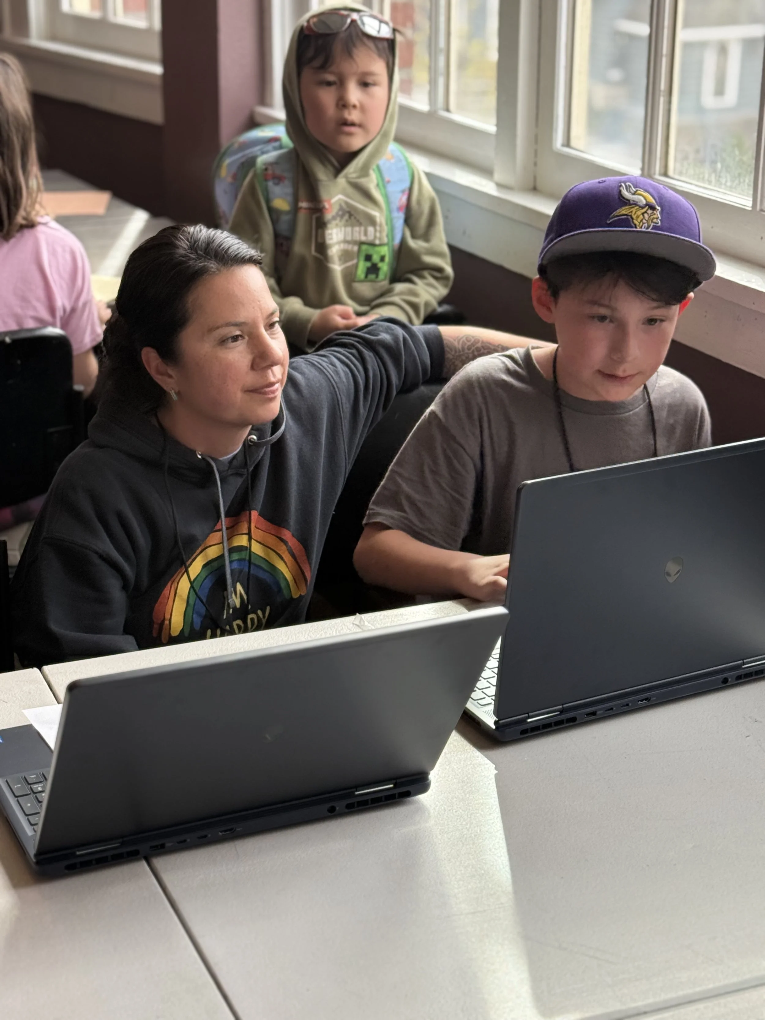 A woman sitting at a table with two laptops, one in front of her and another beside her, while a young boy wearing a Minnesota Vikings cap looks at a laptop screen. A kid in a hoodie with a Minecraft Creeper logo and backpack stands nearby, looking a
