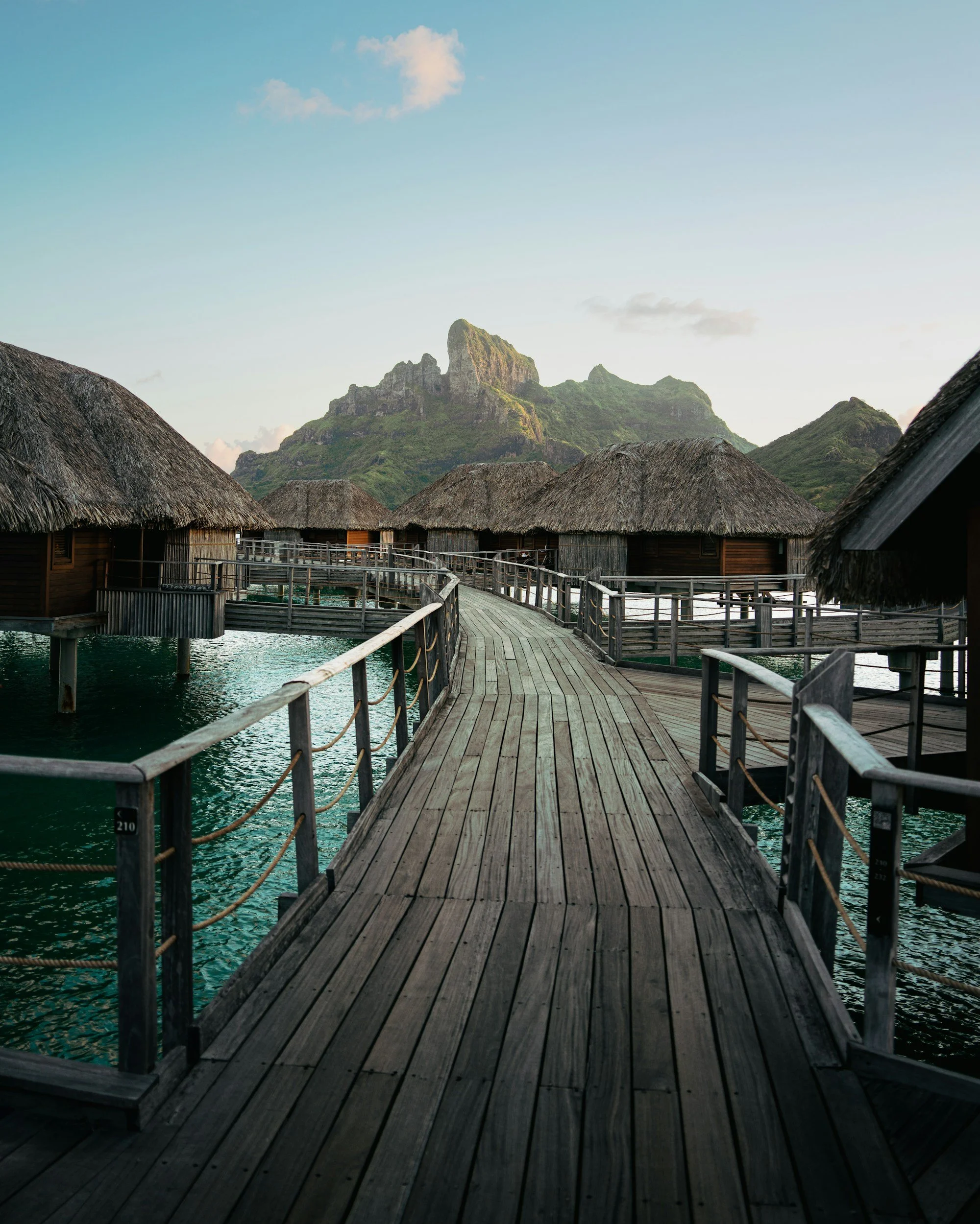 Overwater bungalows with thatched roofs along a wooden pier in a tropical setting with green mountains and blue sky in the background.
