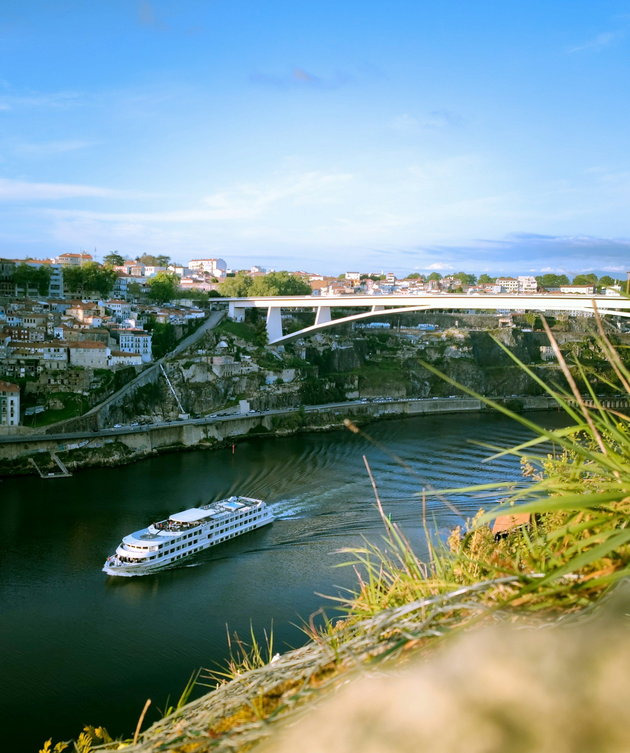 A white cruise boat sailing on a river beneath a bridge, with a hillside filled with houses and trees in the background, and a clear blue sky overhead.