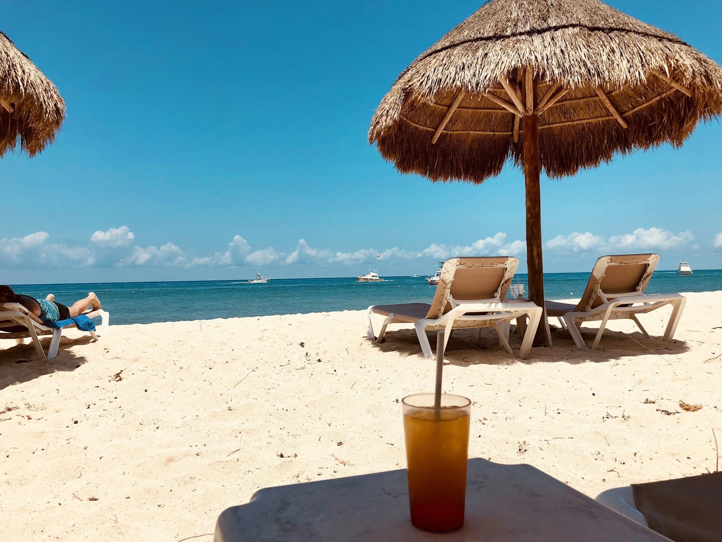 Beach scene with straw umbrellas, lounge chairs, and boats on the water, with a clear blue sky.