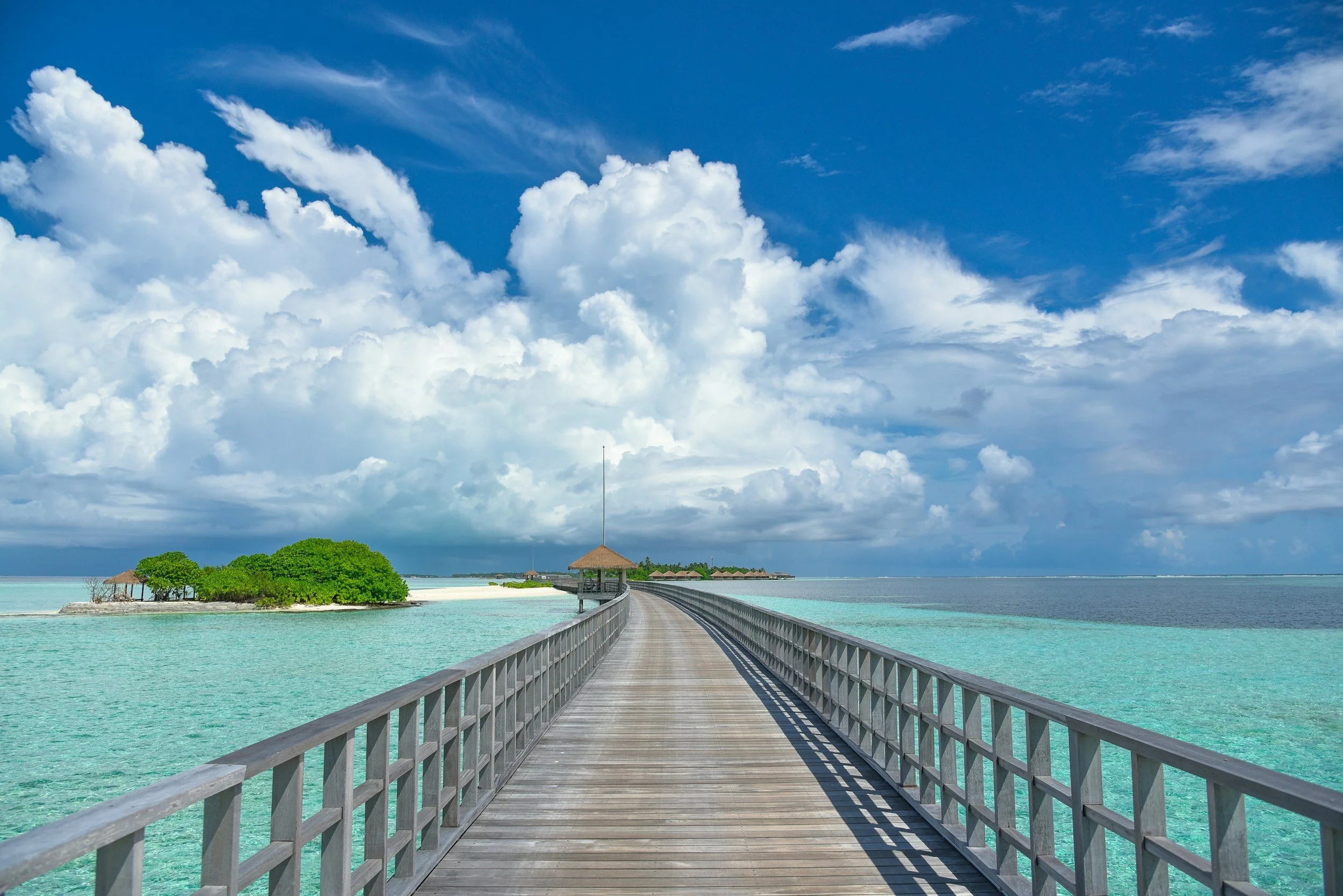 A wooden pier extending over turquoise water towards overwater bavilions and a gazebo, with green foliage on a small island, under a partly cloudy sky.
