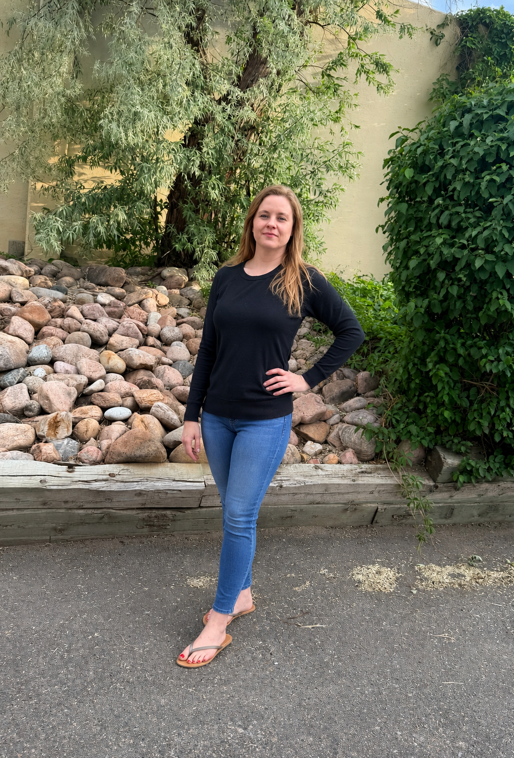 A woman with long blonde hair wearing a black long-sleeve shirt and blue jeans standing outdoors on a sidewalk with her hand on her hip. Behind her is a large rock garden with various-sized rocks and greenery, including trees and bushes.