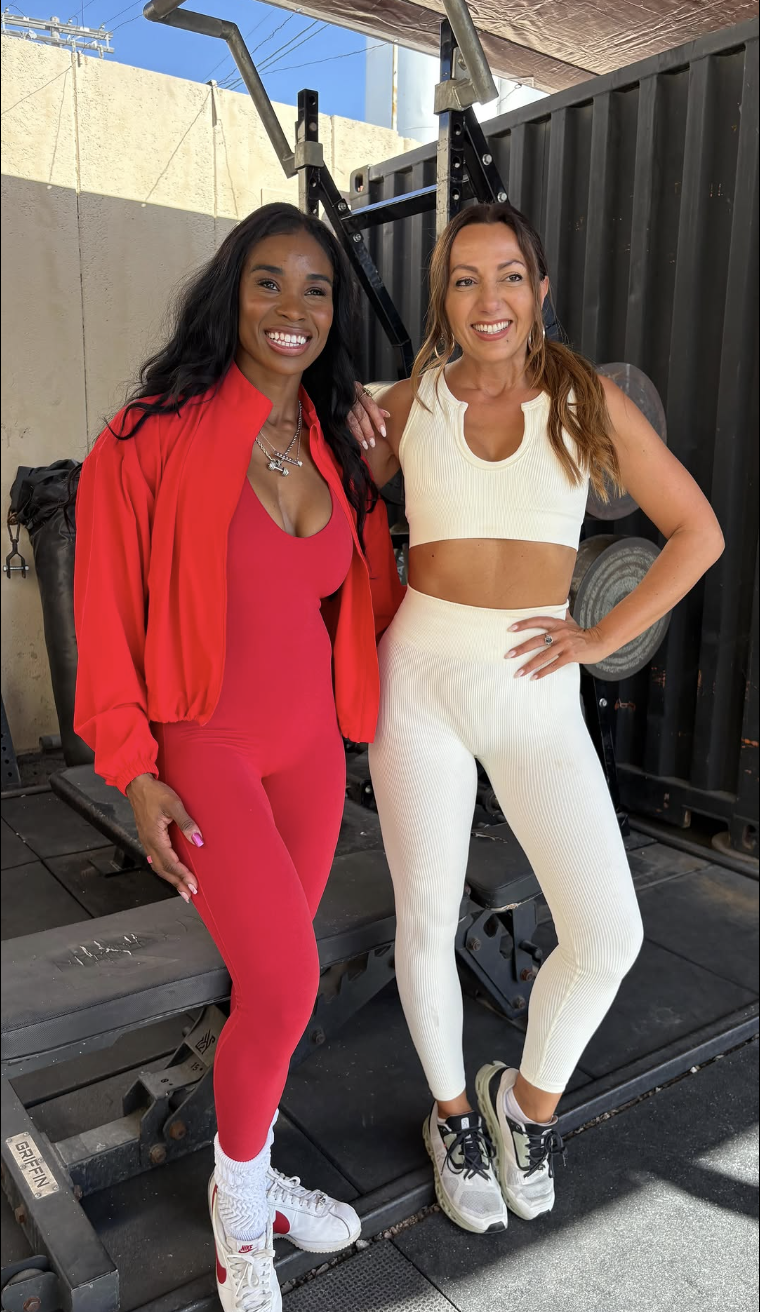 Two women standing in a gym, smiling and posing for the picture. One woman is dressed in red workout clothes, and the other in white workout clothes.