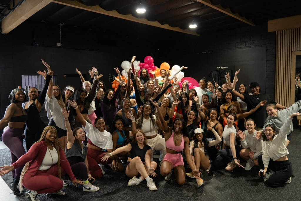 Group of smiling young women and women in athletic clothing celebrating together at in an indoor gym or studio with black walls and balloon decorations.
