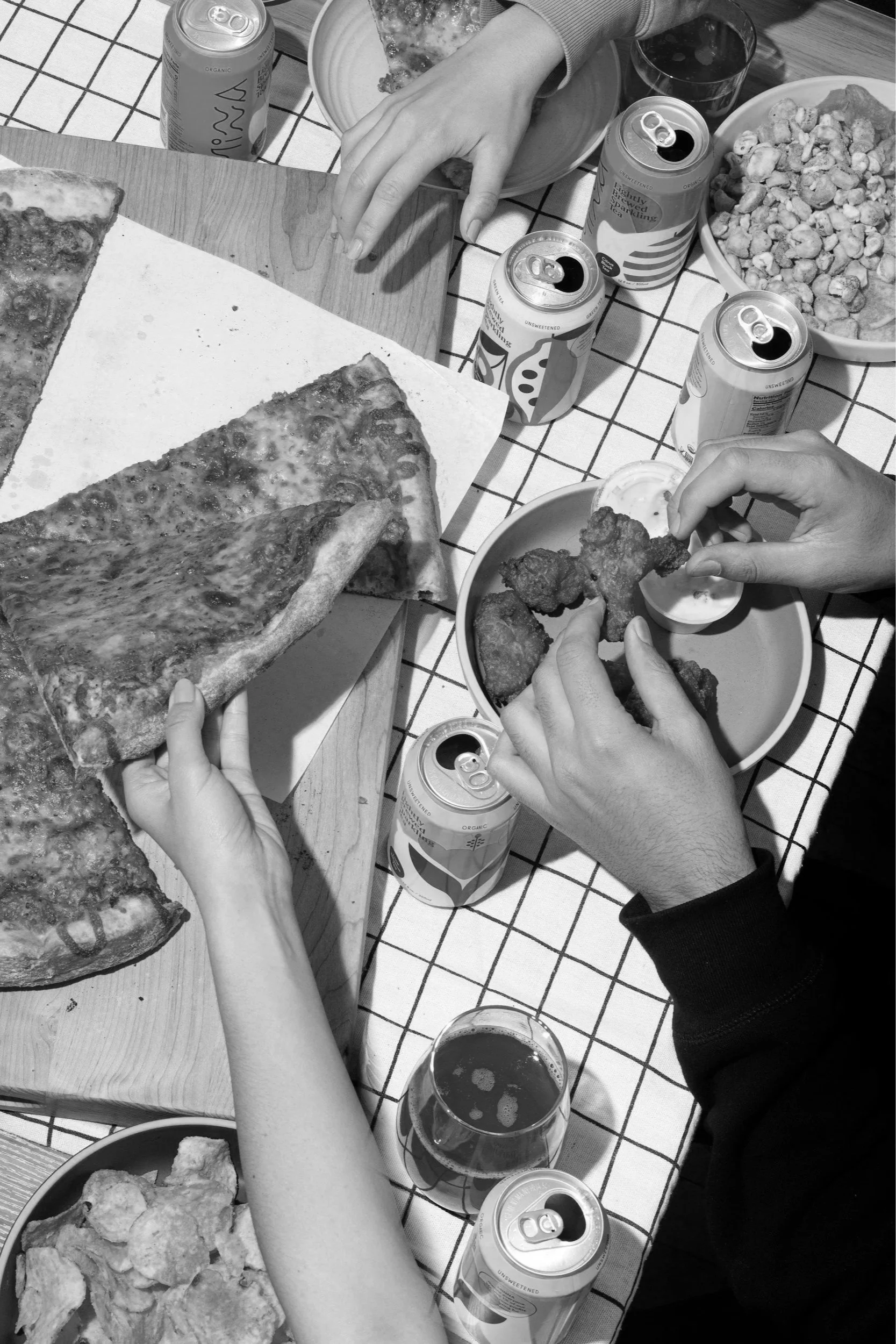 A table with food and drinks, including pizza, fried chicken, soda cans, and glasses of soda, with several people reaching for the food.