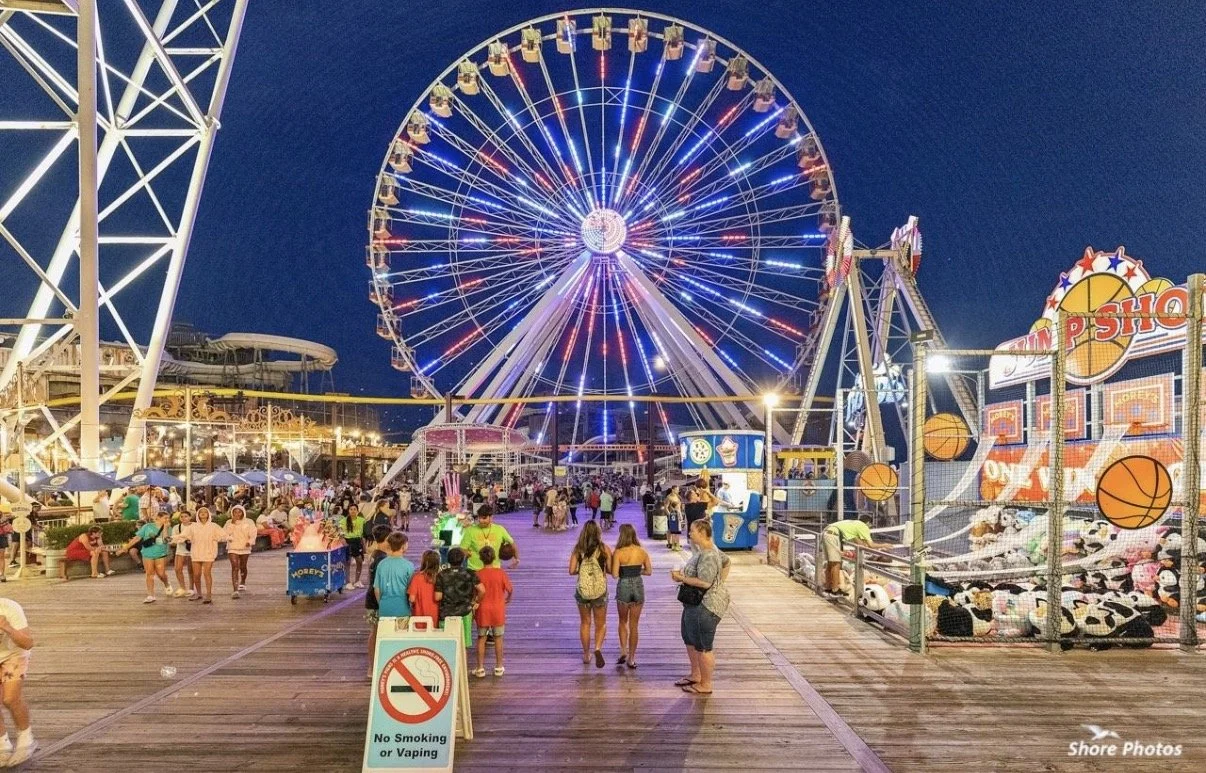People walking on a boardwalk at a carnival with a large illuminated Ferris wheel in the background at night.