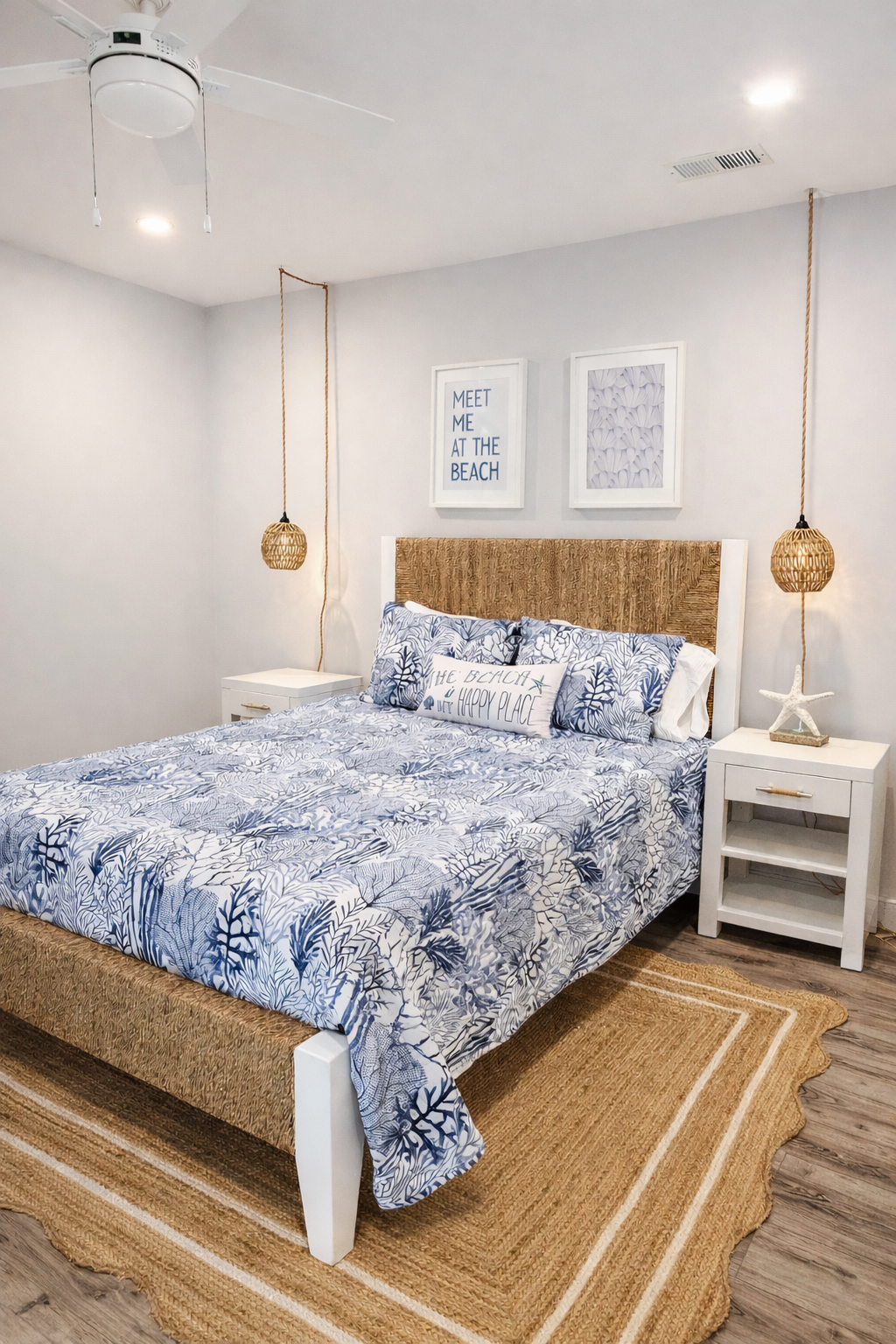 A beach-themed bedroom with a bed featuring a blue and white tropical print bedspread, decorative pillows, and a woven headboard. There are two white nightstands, each with a starfish and woven pendant lights. The wall above the bed has framed beach-themed artwork and a sign that reads "Meet me at the beach." The room has a natural fiber rug and wood flooring.