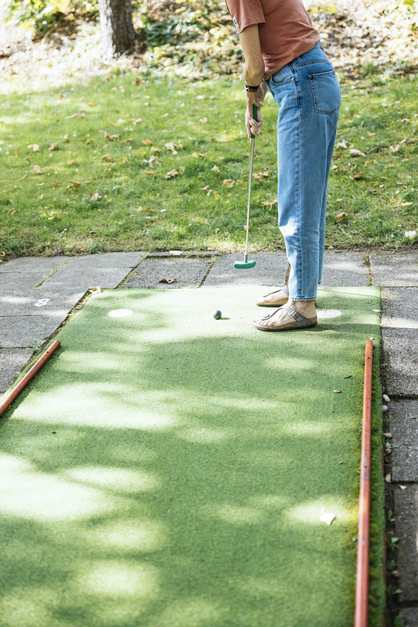 Person playing mini golf on an outdoor course, holding a golf putter and aiming at a white golf ball on a green artificial turf. The scene is set in a park with trees and grass, with outdoor natural lighting.