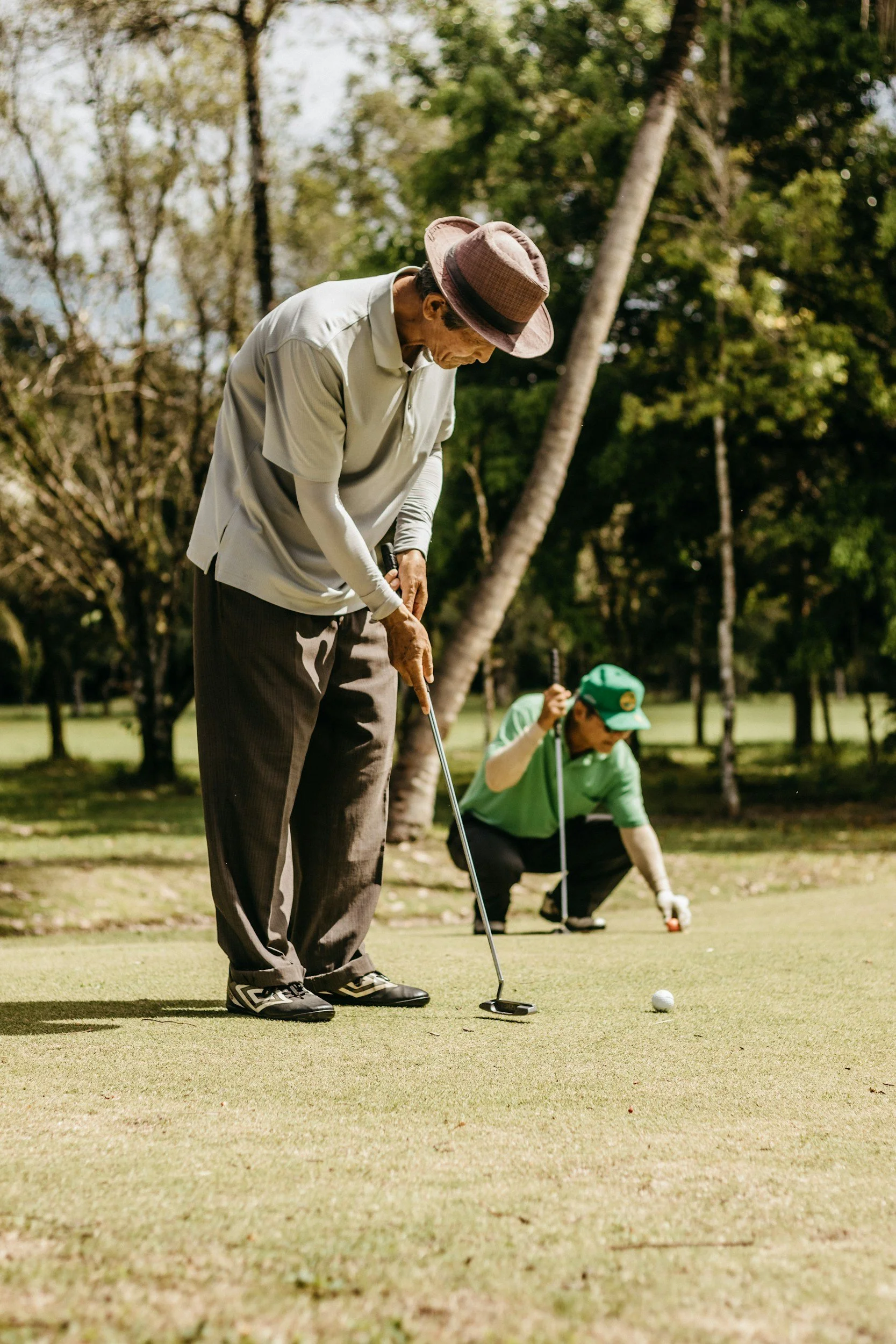 A man in a beige shirt and brown hat lining up a golf putt on a green. A person in a green cap and shirt is crouched nearby, holding a golf club and ball. Trees and grass surround them outdoors.