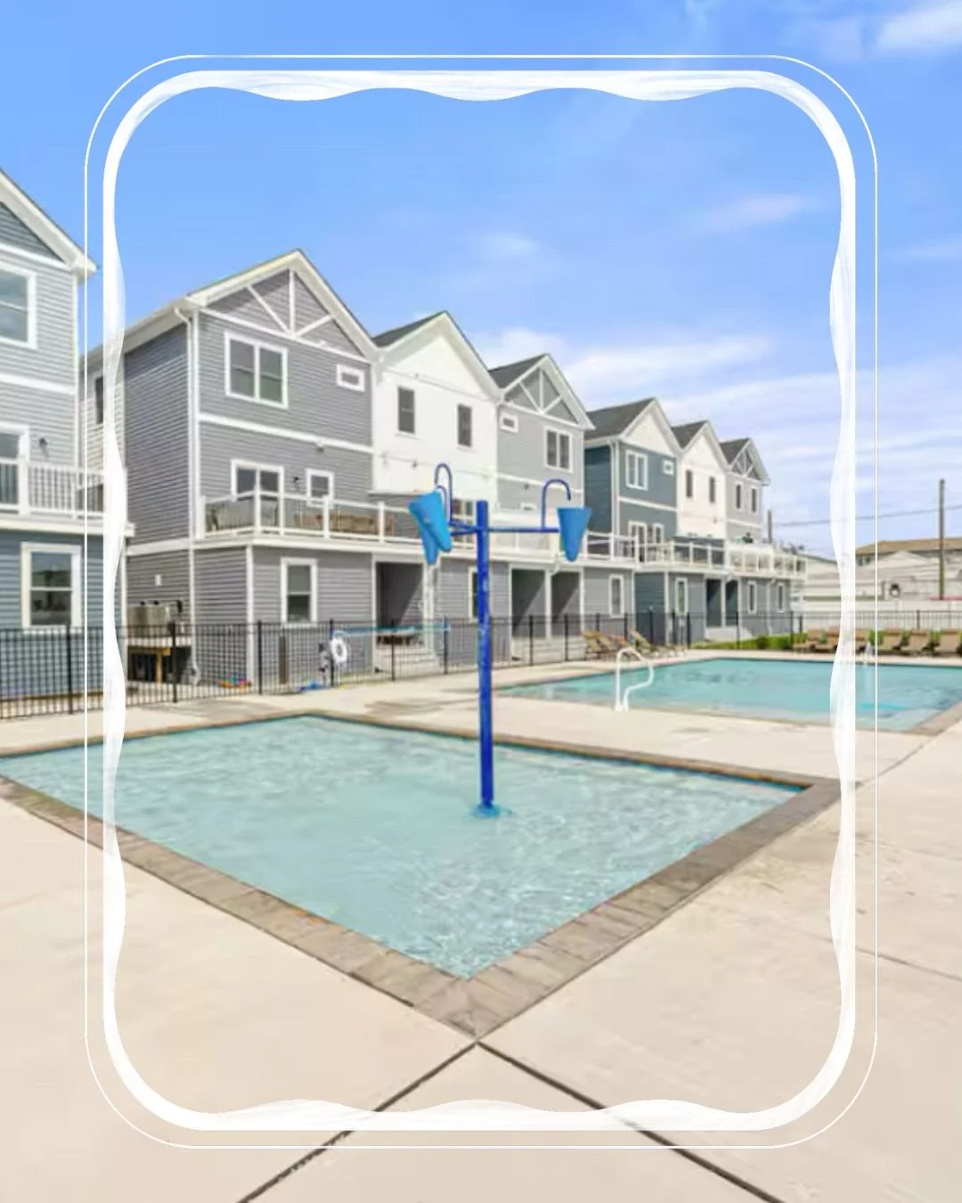 A swimming pool area with a blue water slide and a basketball hoop, surrounded by houses with balconies and a black fence, under a partly cloudy sky.