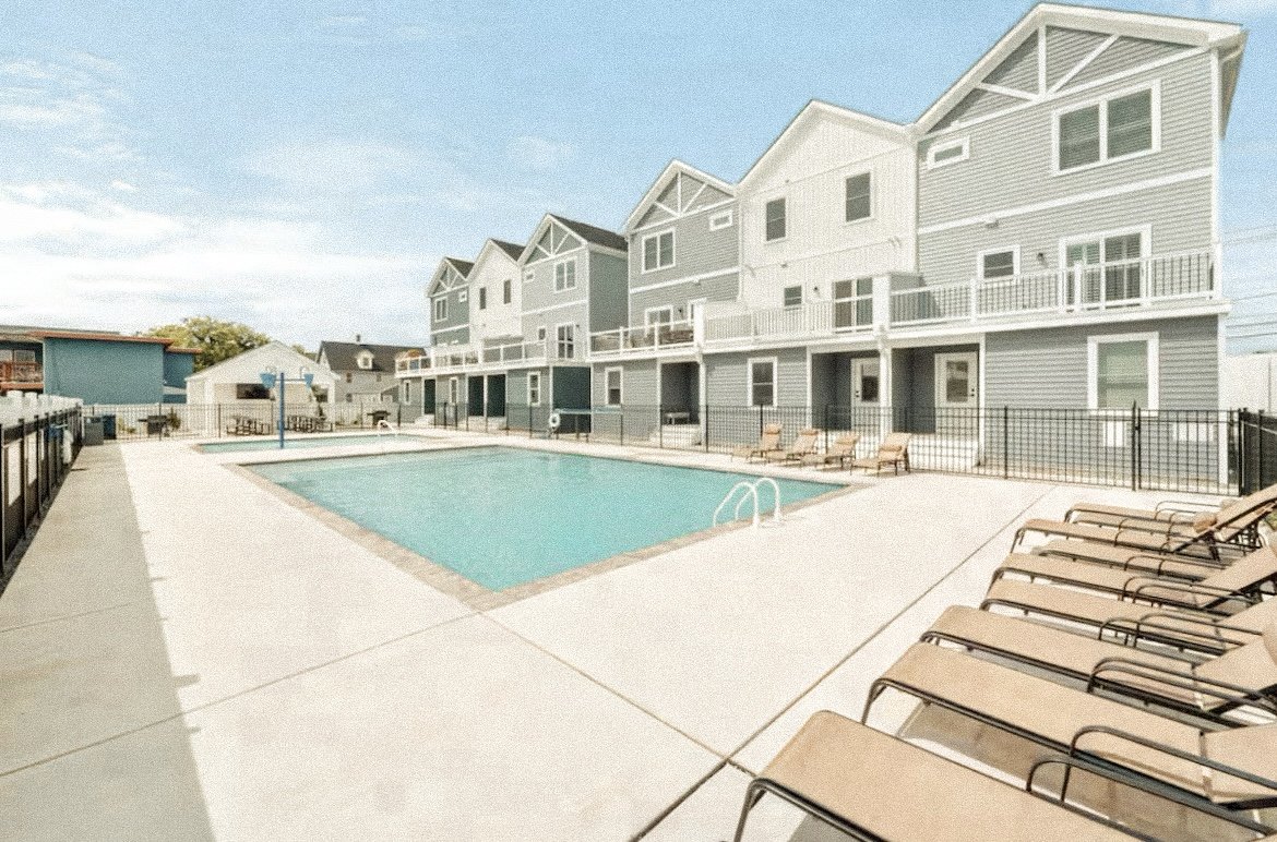 Empty outdoor swimming pool with surrounding lounge chairs, fenced enclosure, and residential buildings in the background under a partly cloudy sky.